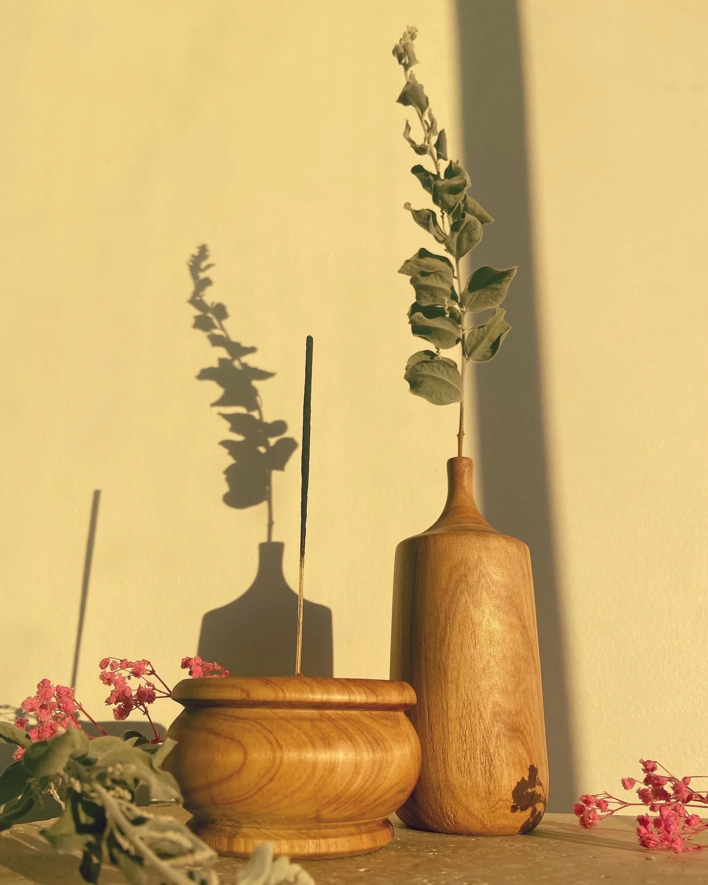 Wooden vases with plants and pink flowers, cast shadows on a yellow wall.