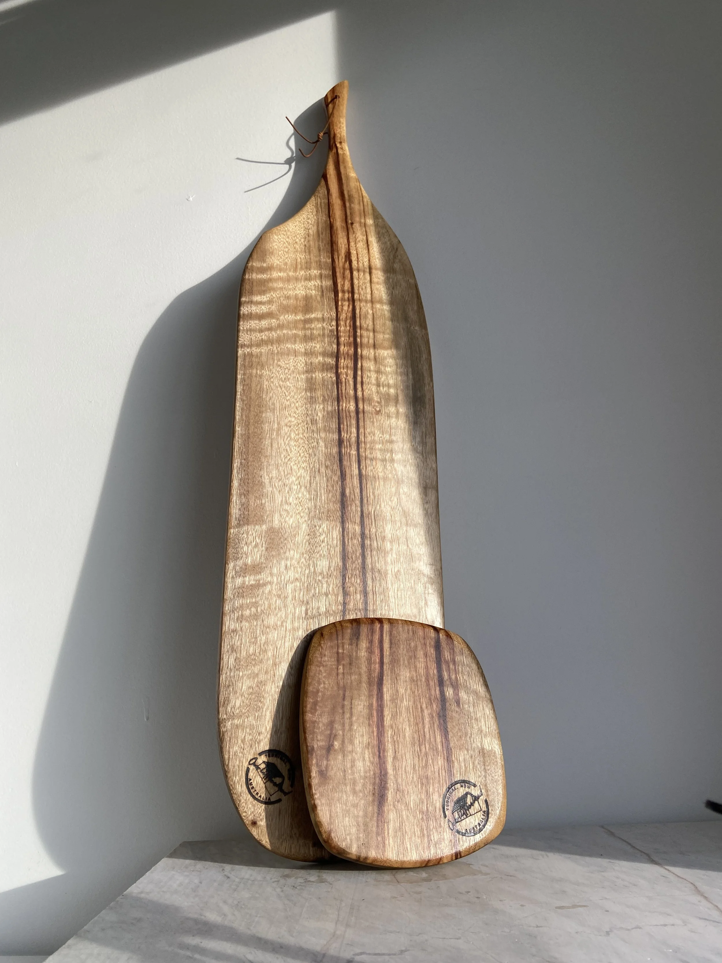 Two wooden cutting boards leaning against a white wall with sunlight casting a shadow, one large and one small, both stamped with a black logo.