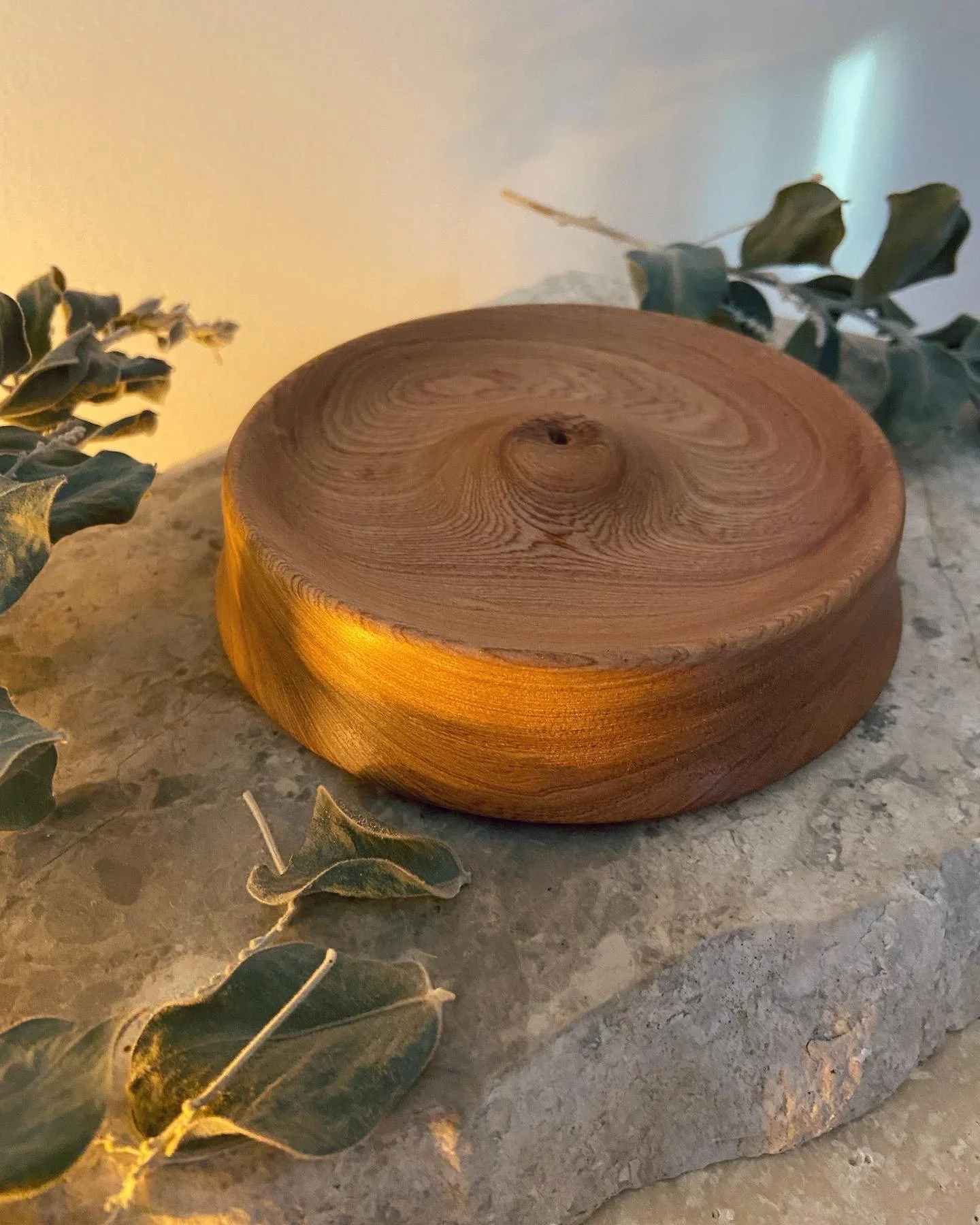 A round wooden container placed on a stone surface with green eucalyptus leaves around it.