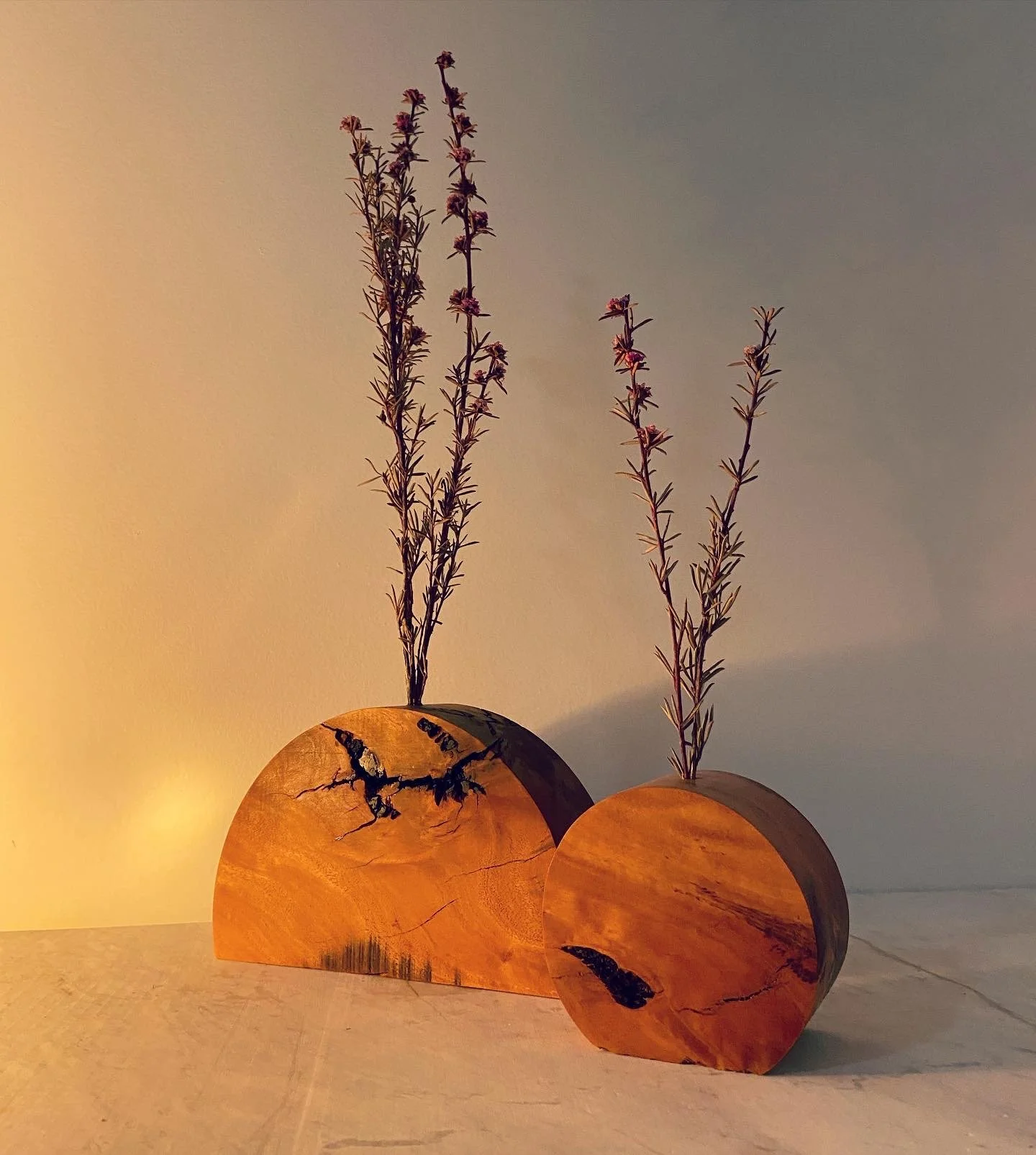 Two wooden vases with dried lavender flowers, one larger and semi-circular, the other smaller and round, placed on a light-colored surface against a plain wall.