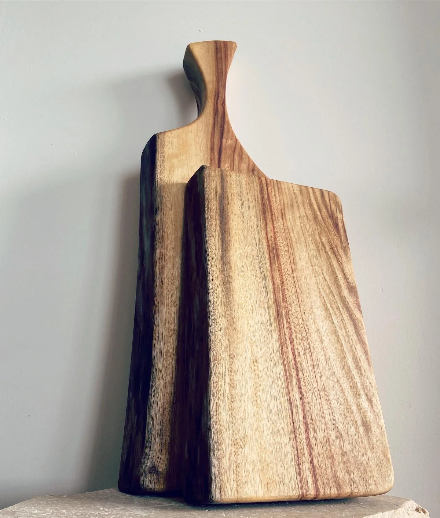 A multi-tiered wooden cutting board with a unique curved handle, made from different wood grains and colors, placed on a light-colored surface against a plain wall.
