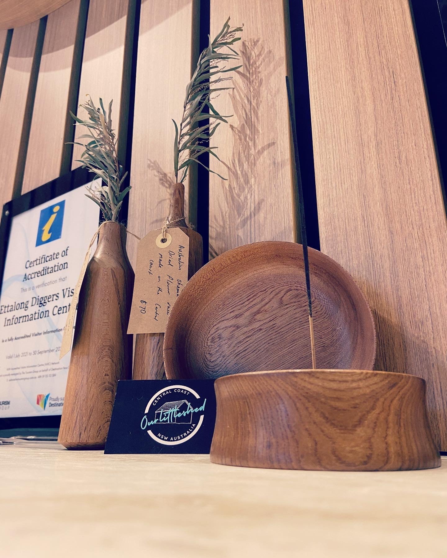 Wooden bowls and dried plant stems arranged on a wooden surface against a wooden wall, with a certificate of accreditation and a small black and white sticker labeled 'Outlanded Central Coast NSW Australia'.