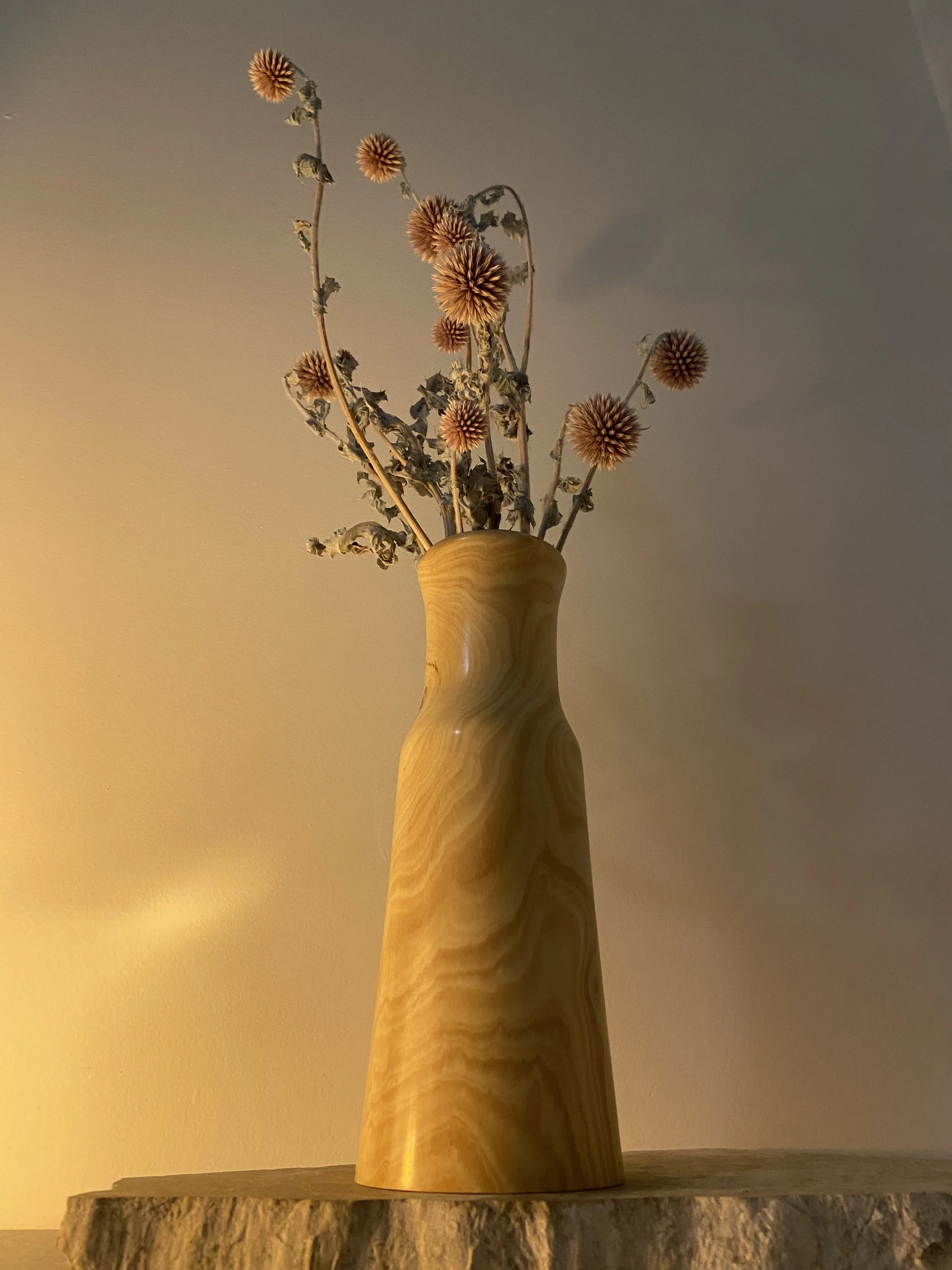 A tall, yellow-marble ceramic vase with a wavy design, holding dried pinkish-orange spherical flowers and plant stems, placed on a wooden surface against a plain wall.