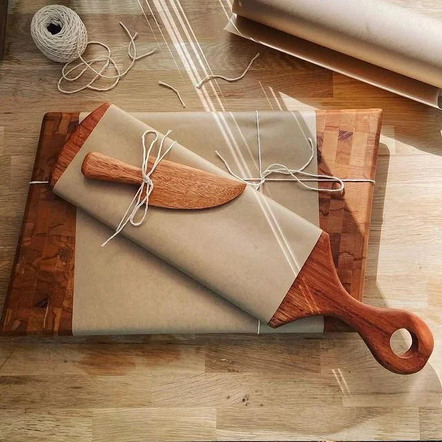 A wooden cutting board with rolled parchment paper tied with string, a wooden dough scraper, additional parchment paper, and a ball of twine on a light wooden surface.