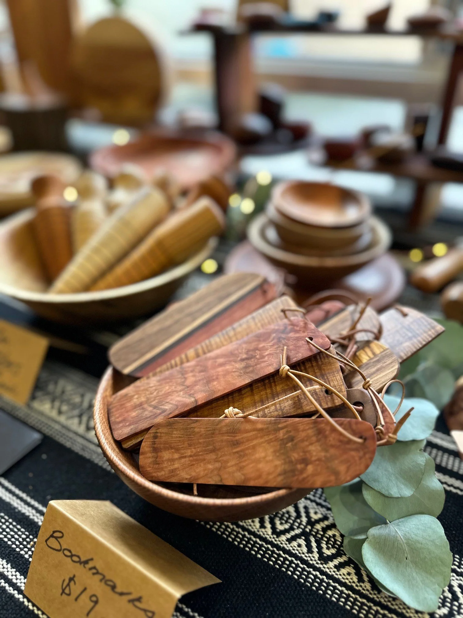 Wooden camping utensils in a bowl with a yellow price tag of $19, displayed on a patterned cloth at a craft market.