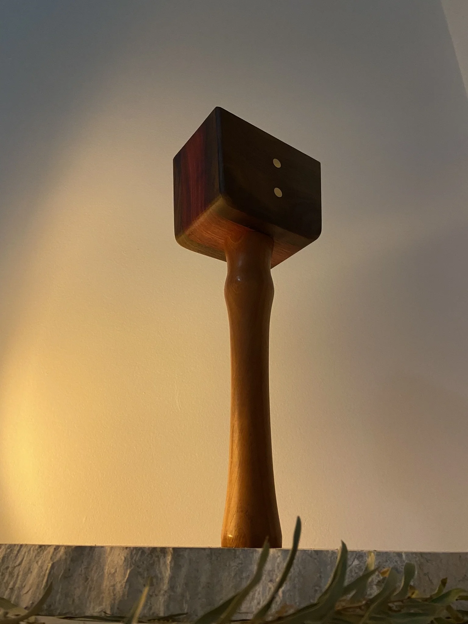 A decorative wooden sculpture resembling a hammer with a cube-shaped head, mounted on a granite base, positioned indoors with a neutral wall background.
