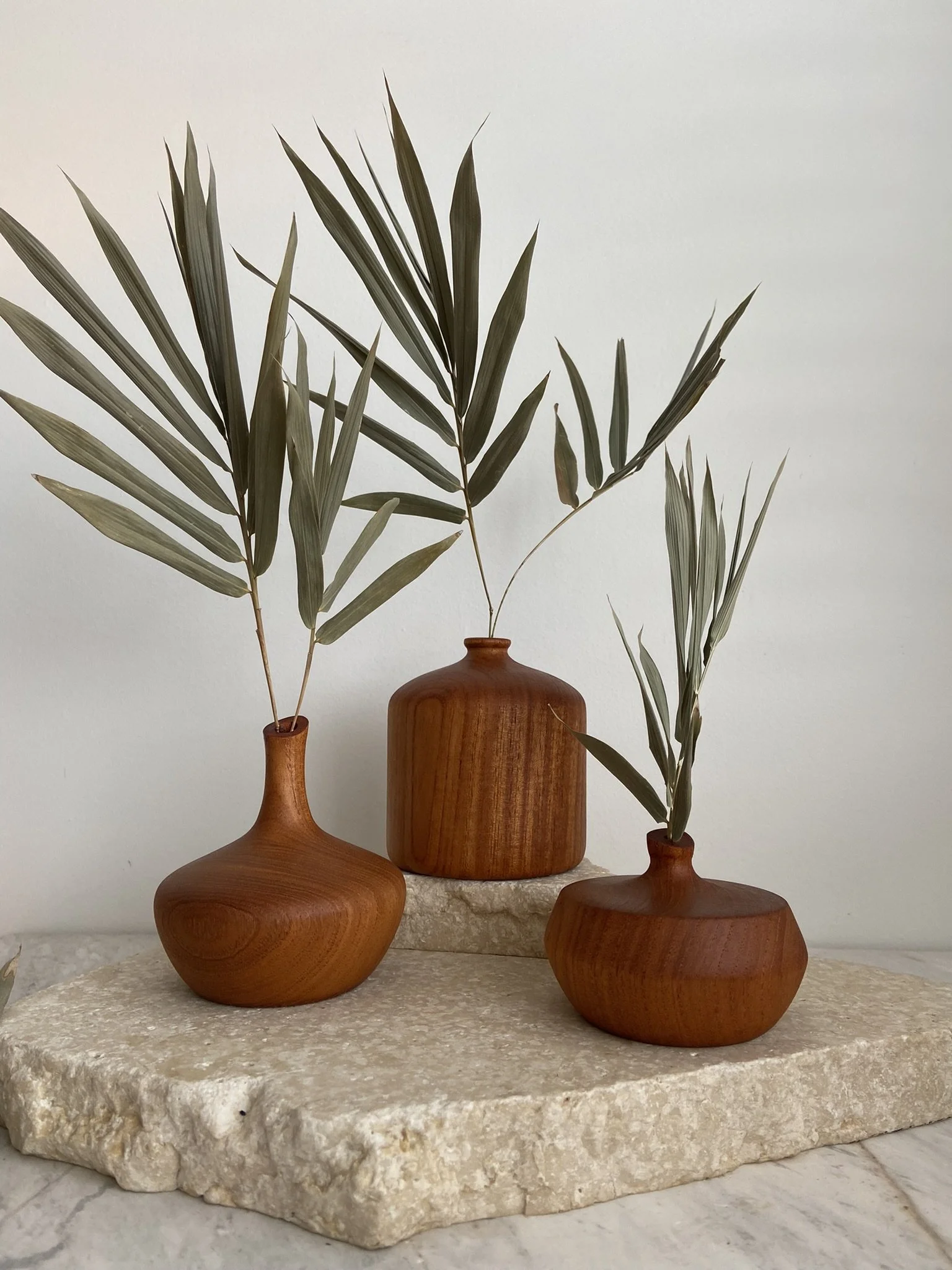 Three wooden vases with long green leaves arranged on a textured beige stone slab against a plain white wall.