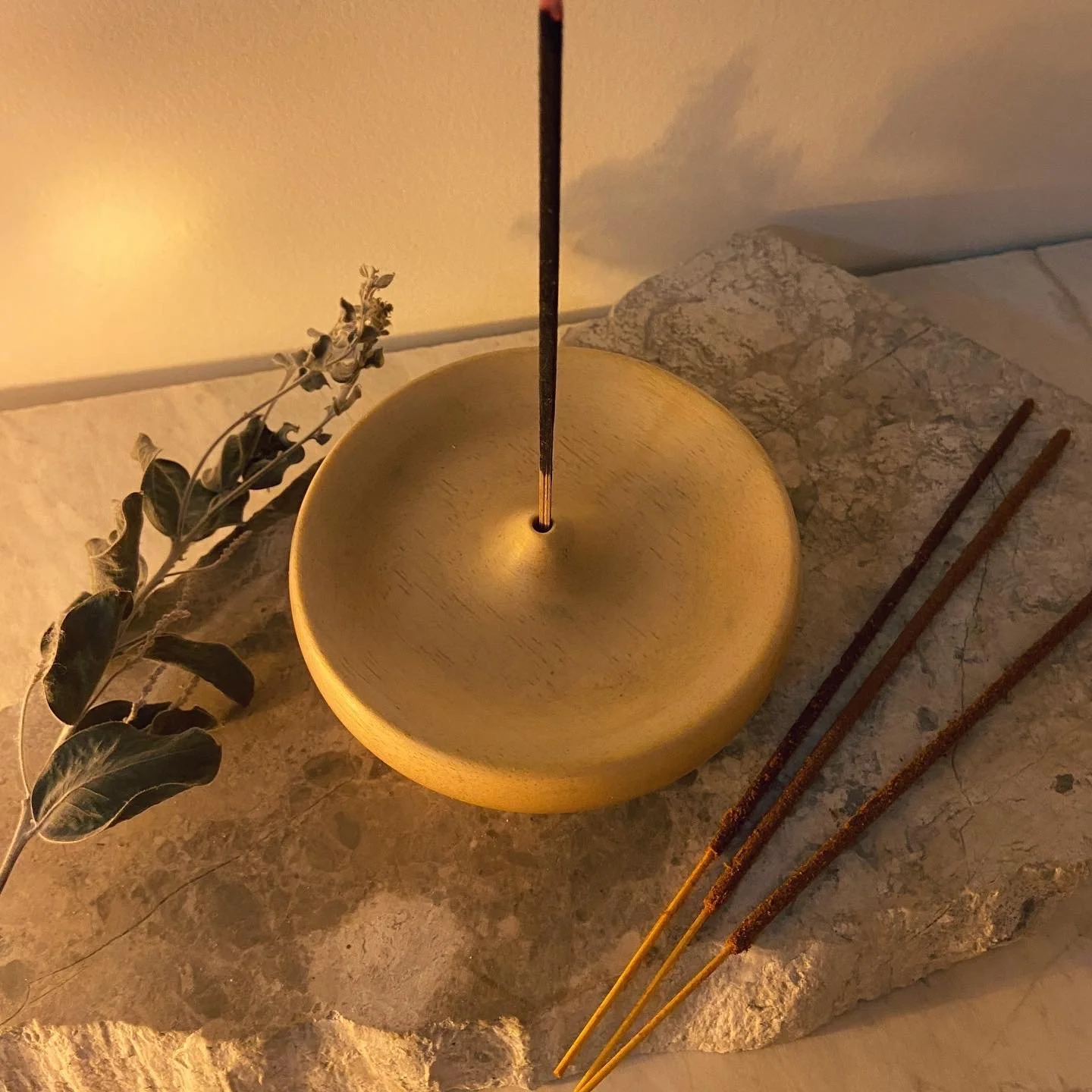 A yellow lantern with a lit incense stick inside, placed on a marble surface with three more incense sticks beside it and a sprig of dried leaves nearby.