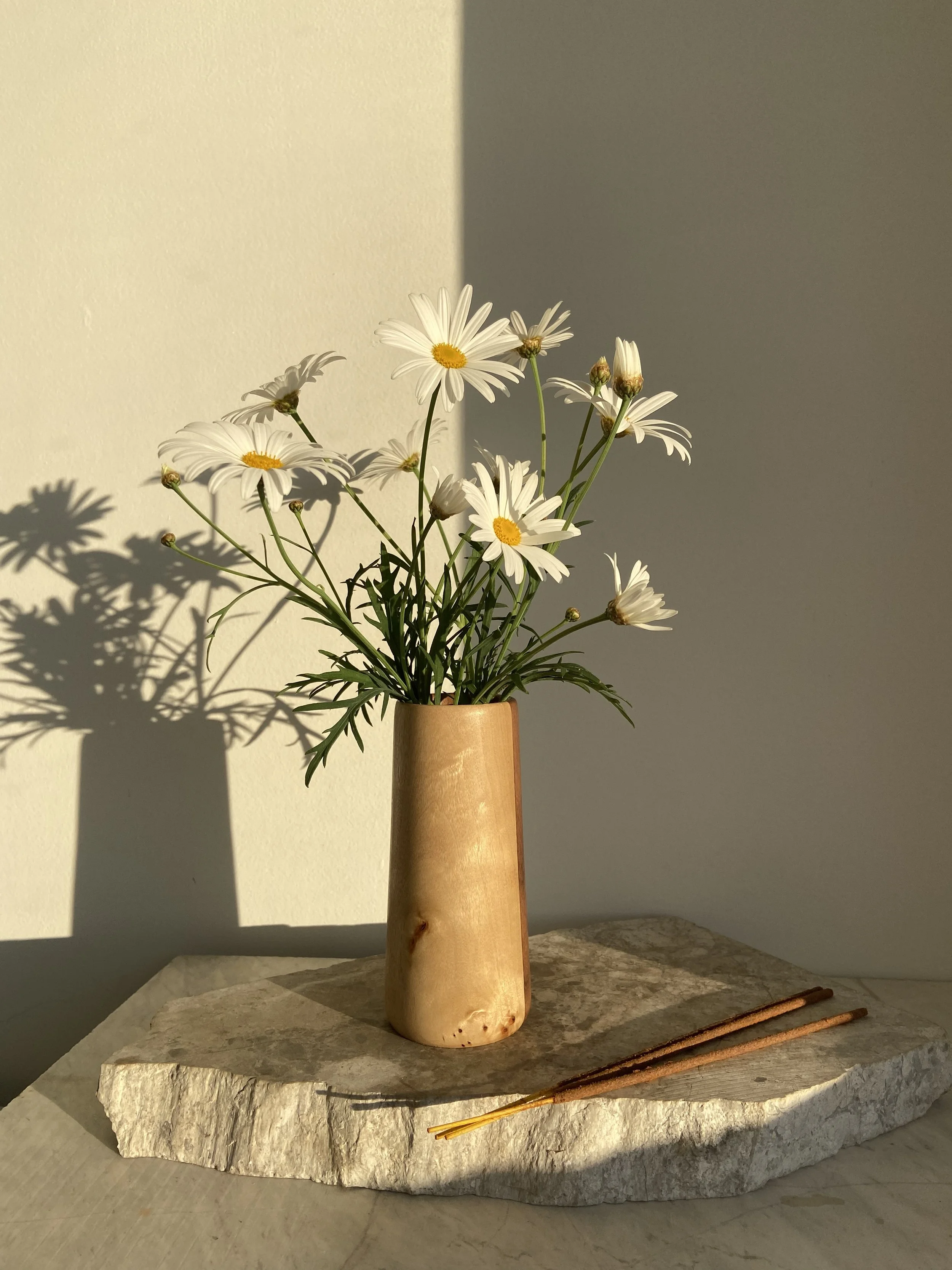 A wooden vase with white daisies on green stems, placed on a textured stone slab, with shadows cast on a plain wall behind.