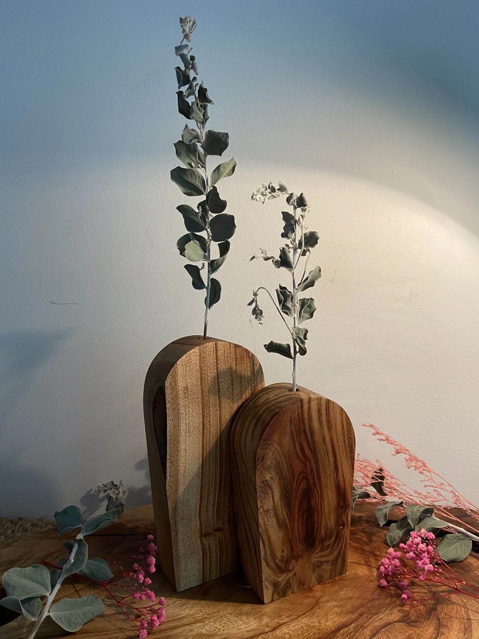 Two wooden vases with green and white foliage, placed on a wooden surface with pink flowers and dried leaves around them.