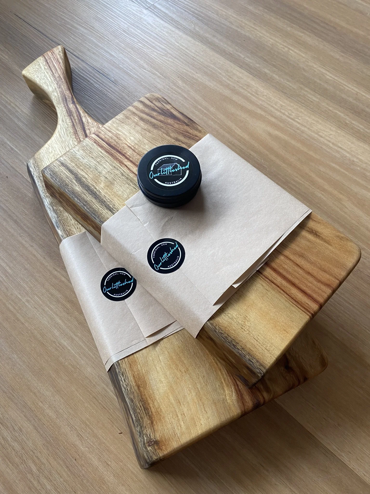 A wooden cutting board with three brown paper bags and three circular stickers, placed on a wooden surface.