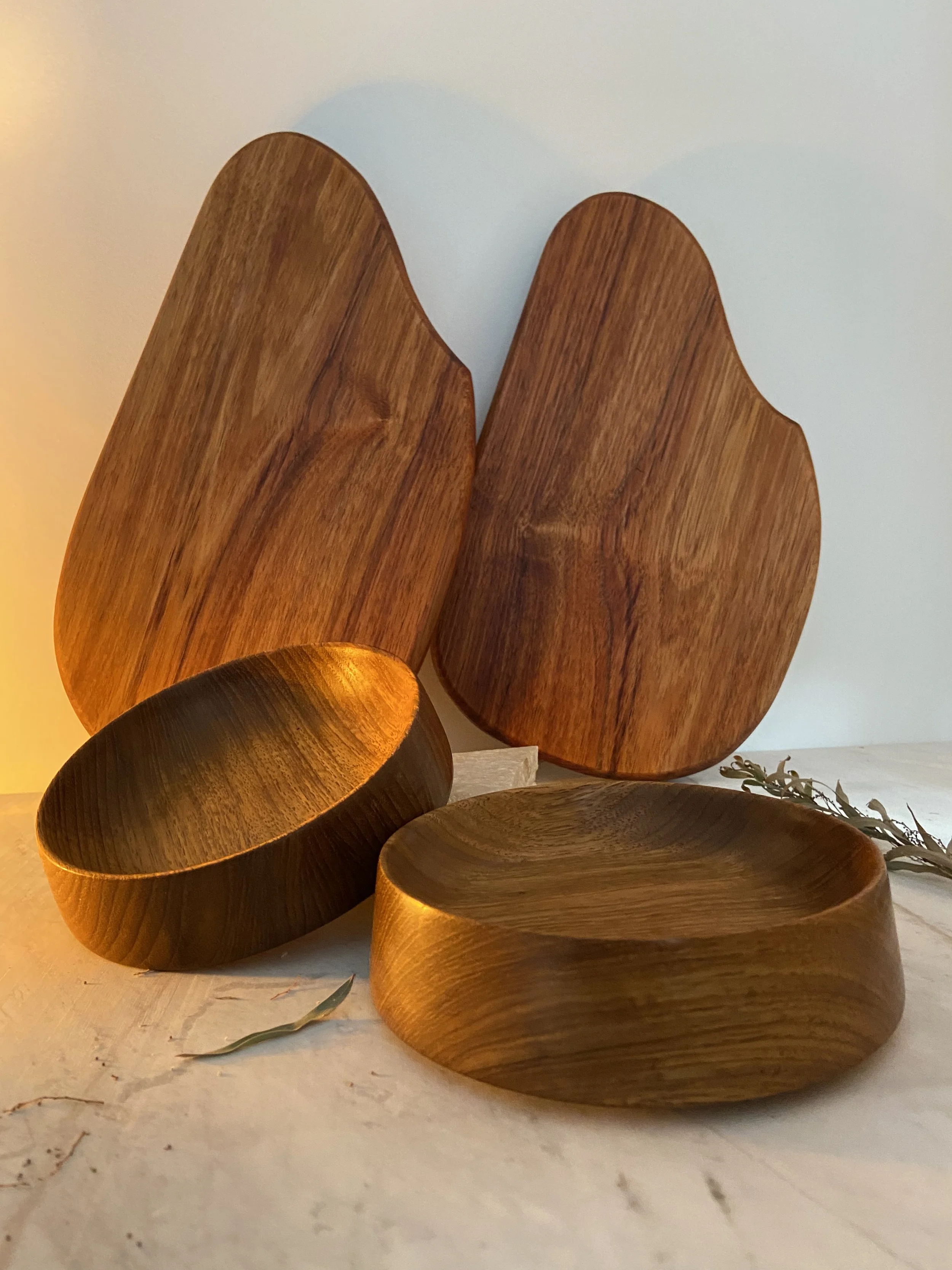 Two large wooden serving boards stand upright in the background, with two round wooden bowls in the foreground, all made of dark polished wood, with a warm lighting casting a soft glow.