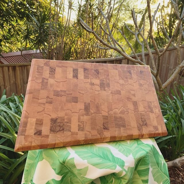 A wooden cutting board resting on a green foliage-patterned cloth outdoors, with trees and a fence in the background.