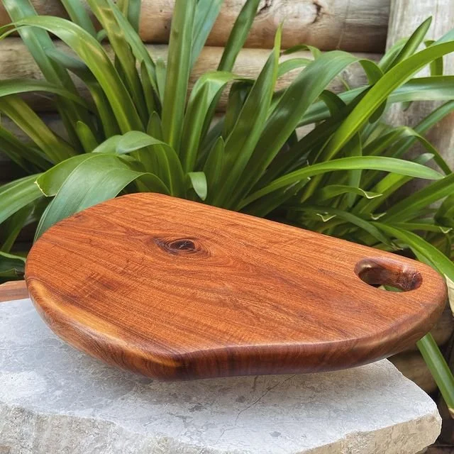 A wooden cutting board with a smooth, polished surface and a handle, placed on a stone surface in front of green leafy plants.