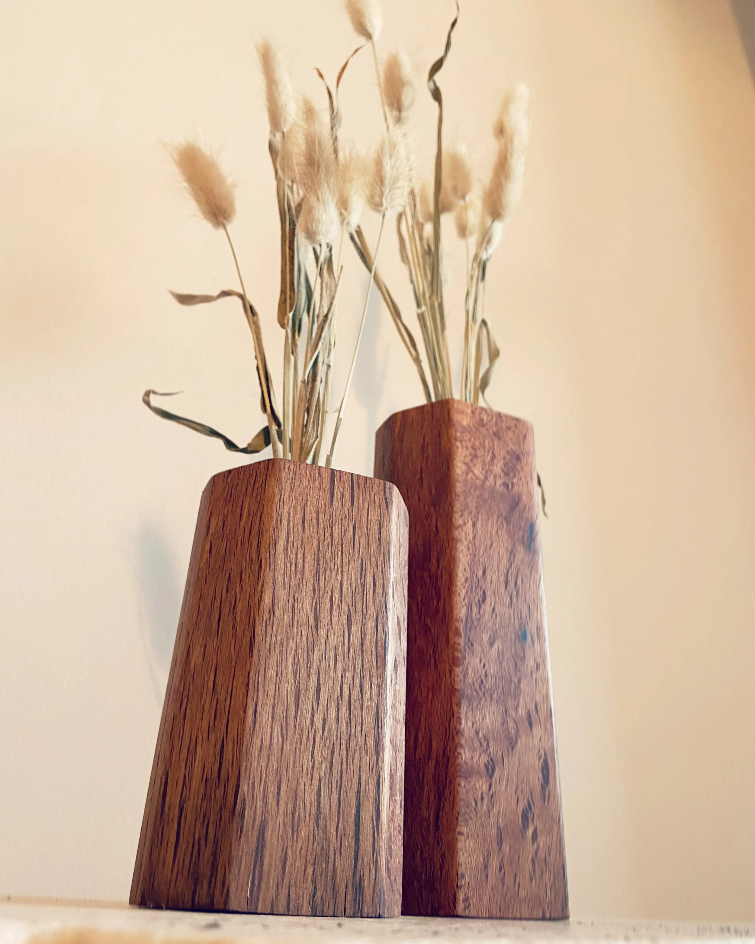 Two wooden vases with dried pampas grass. One vase is shorter and lighter, the other taller and darker. The vases are placed on a flat surface against a plain wall.