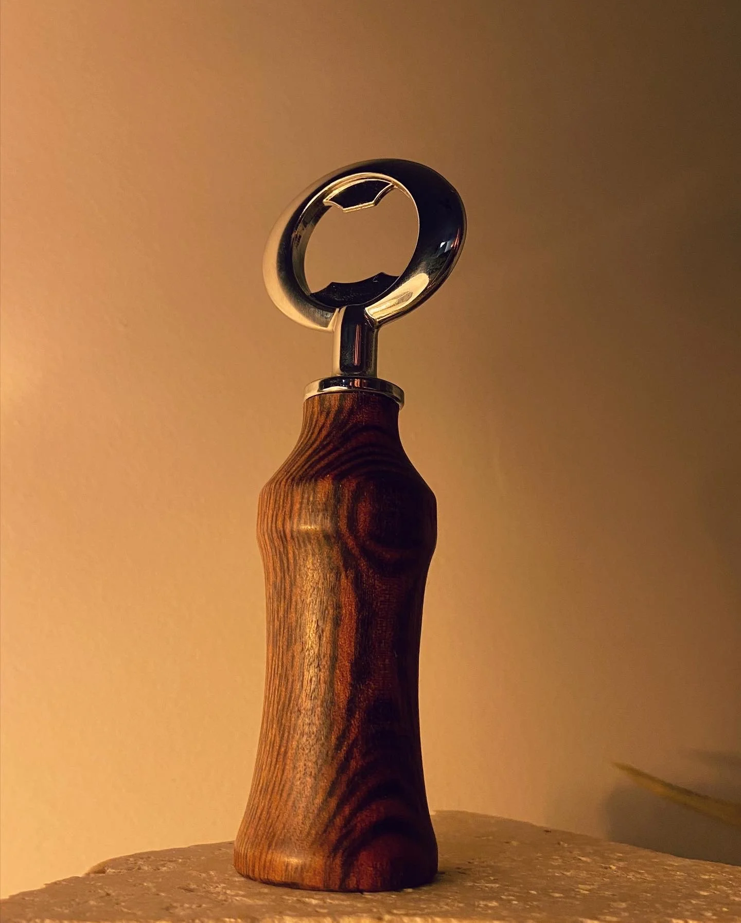 A corkscrew with a wooden handle and a metal screw standing upright on a surface against a warm-colored wall.