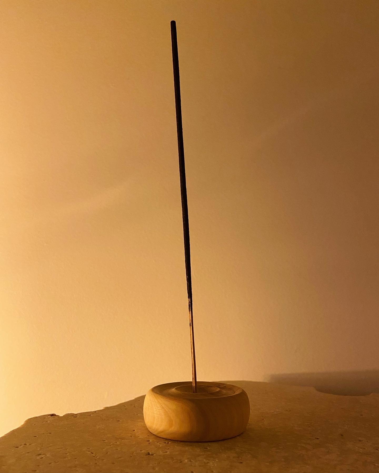 A wooden incense holder with a long, thin incense stick inserted, placed on a wooden surface, illuminated by warm lighting.