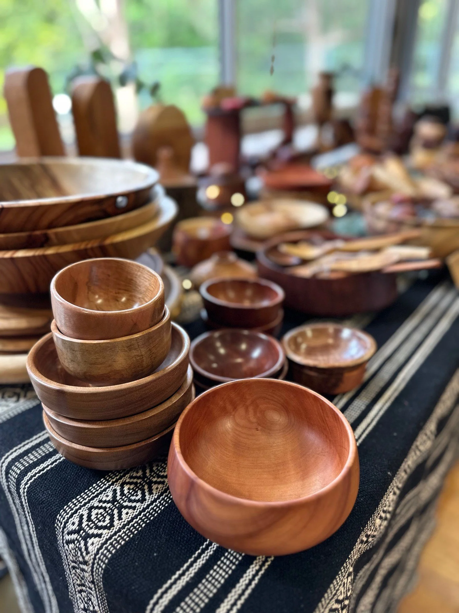 Stack of wooden bowls on a patterned cloth table, with more bowls and wooden items in the background.