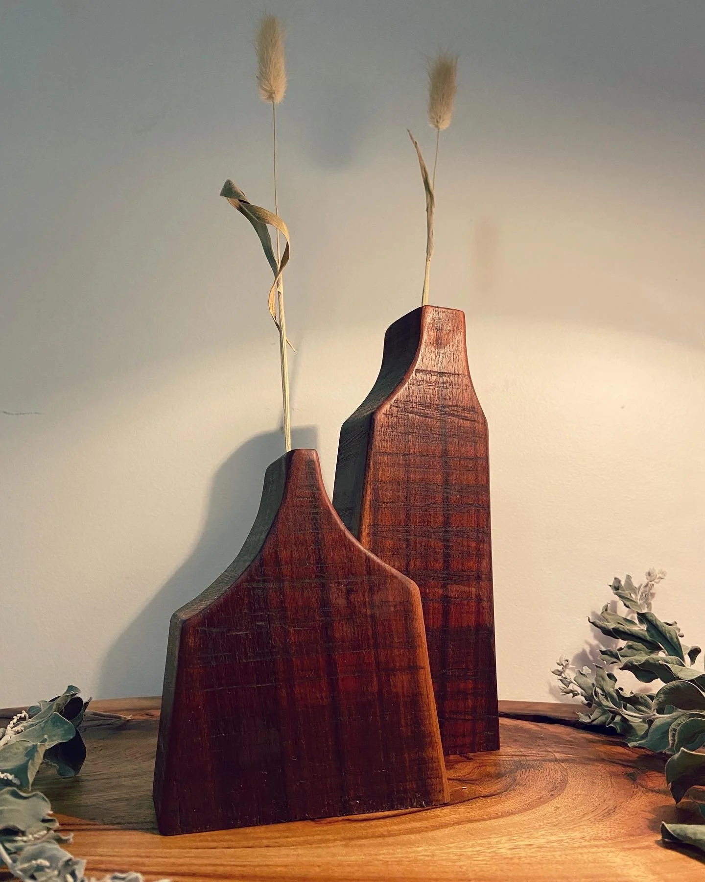Two wooden vases with dried grass stems on a wooden surface, with greenery on the sides.