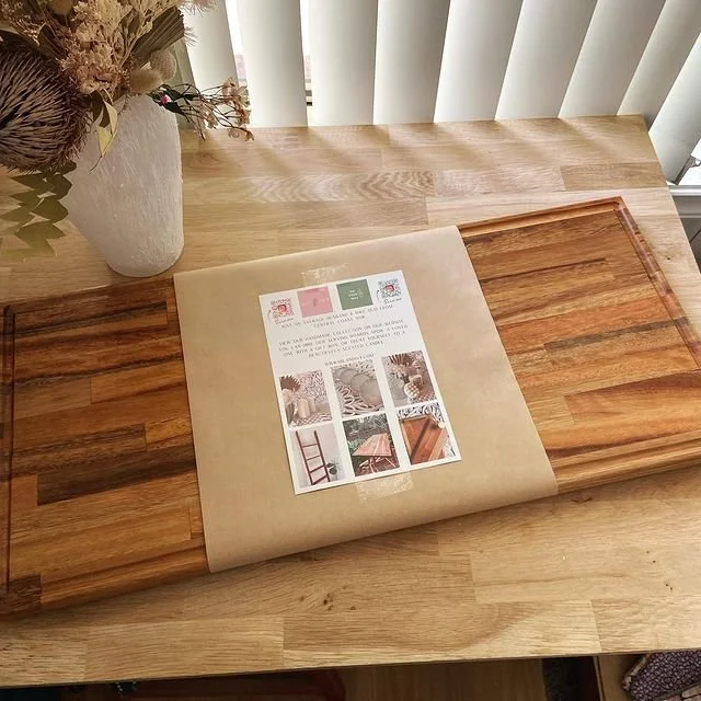 Wooden cutting board with a package on top, placed on a light wood table near a white vase with dried flowers, with vertical blinds in the background.