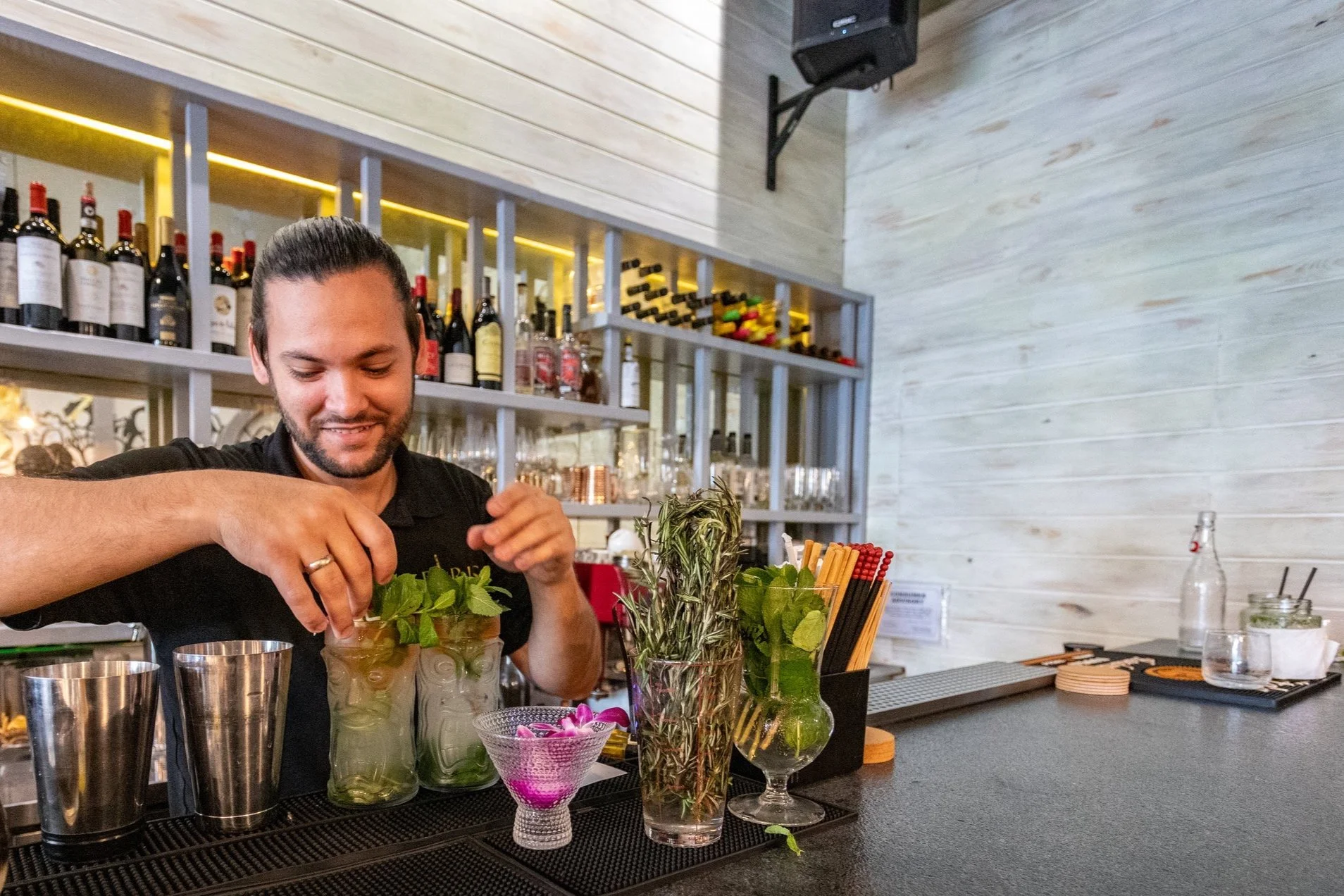 Bartender preparing cocktails with mint and stirring sticks on a bar counter. Background features shelves with various liquor bottles and glassware.