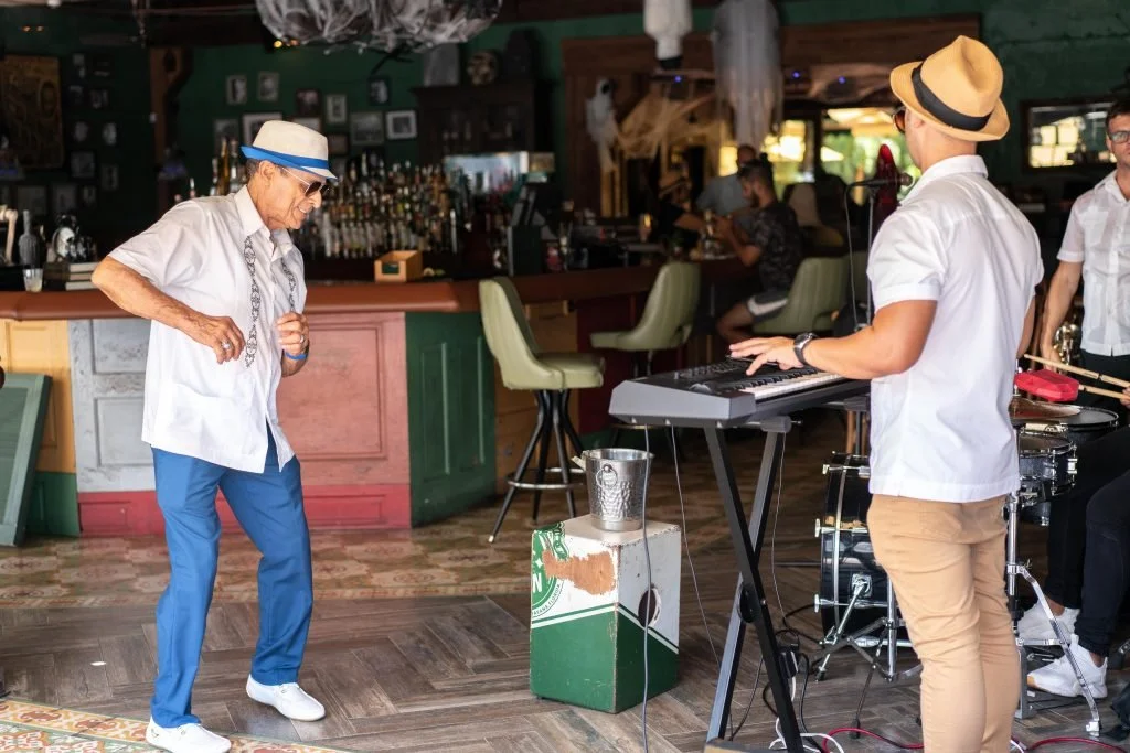 An elderly man wearing a white shirt, blue pants, and a fedora hat dancing in a bar, with a musician playing a keyboard next to drums in the background. The bar is decorated with green walls and a wooden counter.