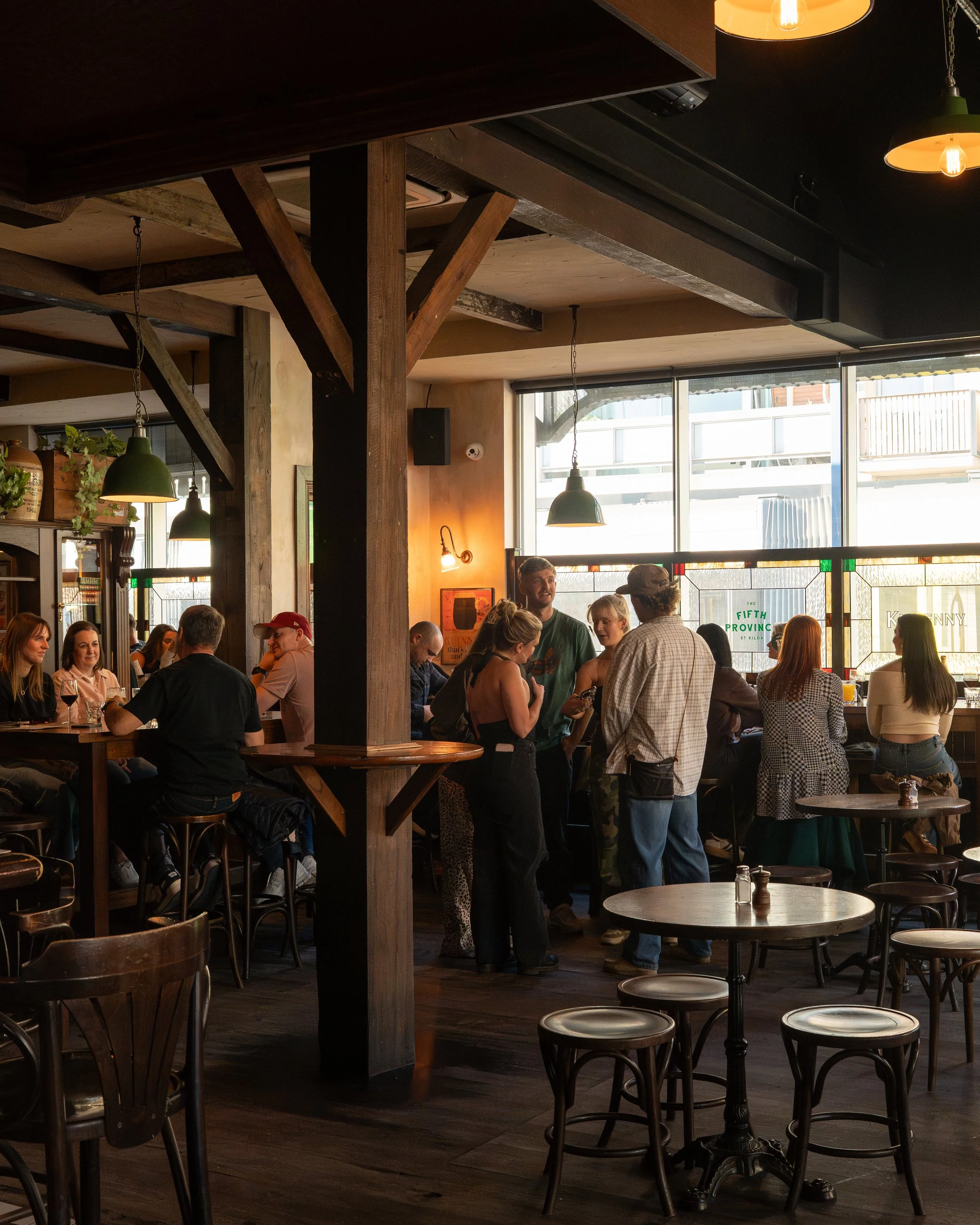 People socializing inside a cozy bar or cafe with wooden furniture, hanging lights, and large windows letting in natural light.