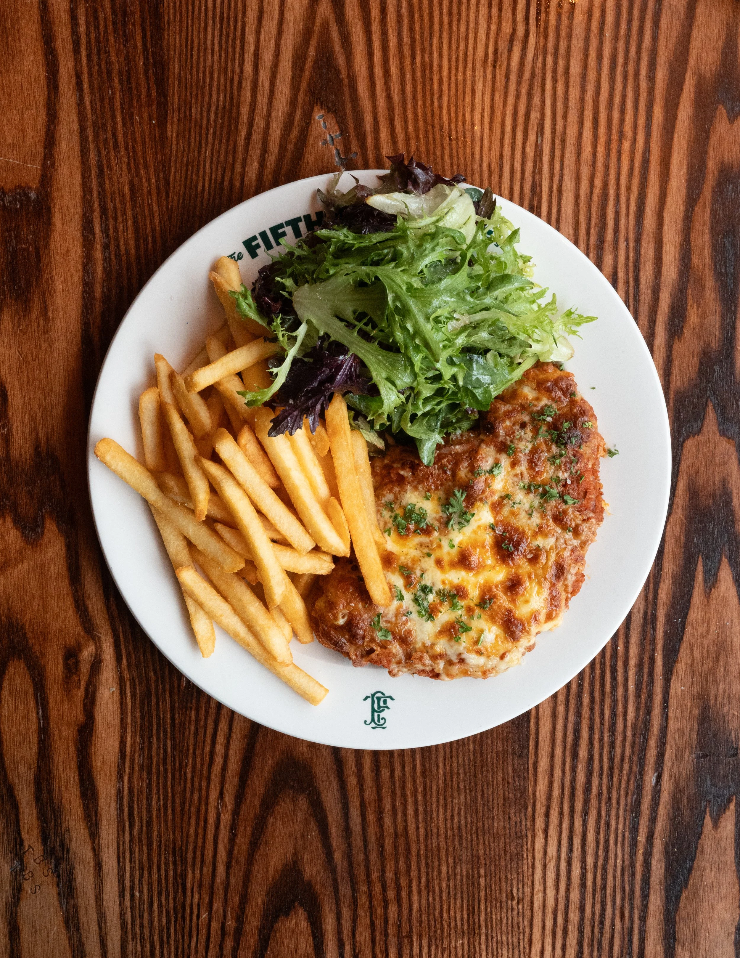 A Chicken Parma topped with cheese, french fries, and a side salad with mixed greens on a wooden table, with two glasses of beer at The Fifth Province.