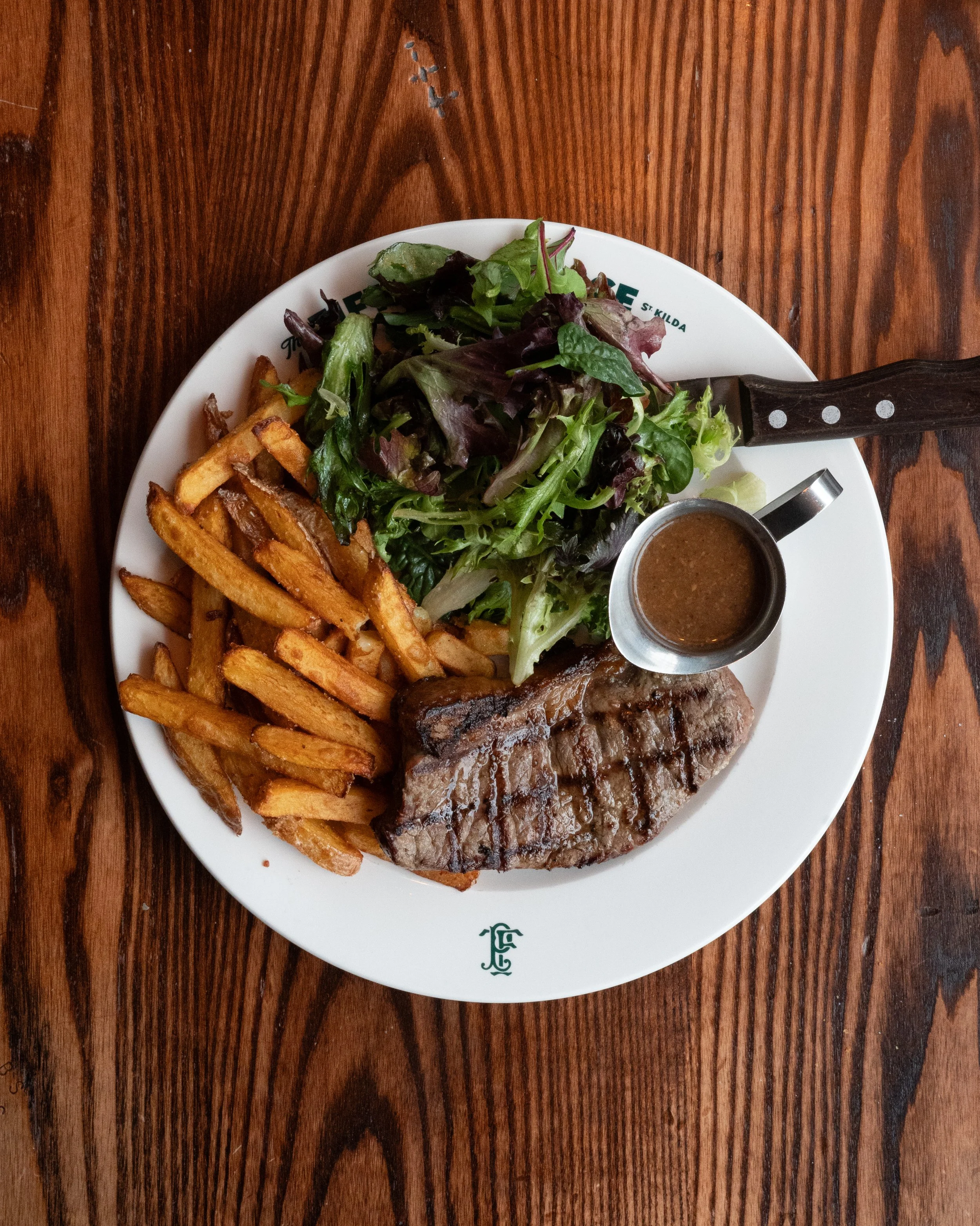A plate with grilled steak drizzled with sauce, French fries, mixed greens salad, and a small container of dark sauce, on a wooden table.
