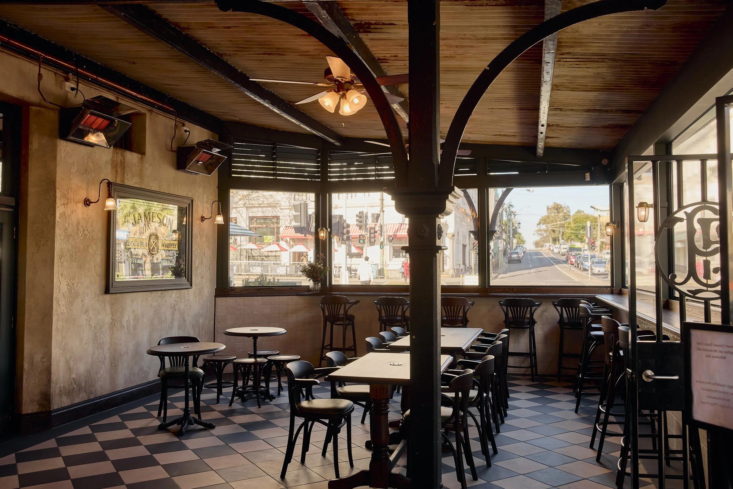 Interior of a cozy restaurant or café with a checkered black and white floor, tables and chairs, large windows letting in natural light, a ceiling fan, and decorative lighting fixtures. Outside, a street scene with buildings, trees, and cars is visible through the windows.