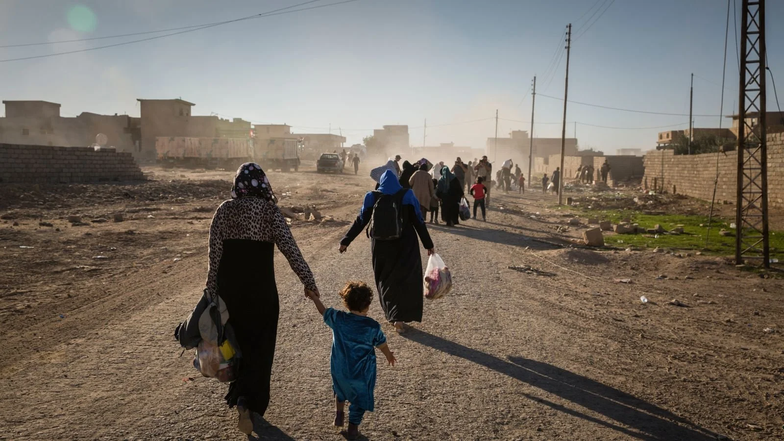 People, including women and children, walking along a dusty, unpaved street in a developing area, with some buildings and utility poles in the background.