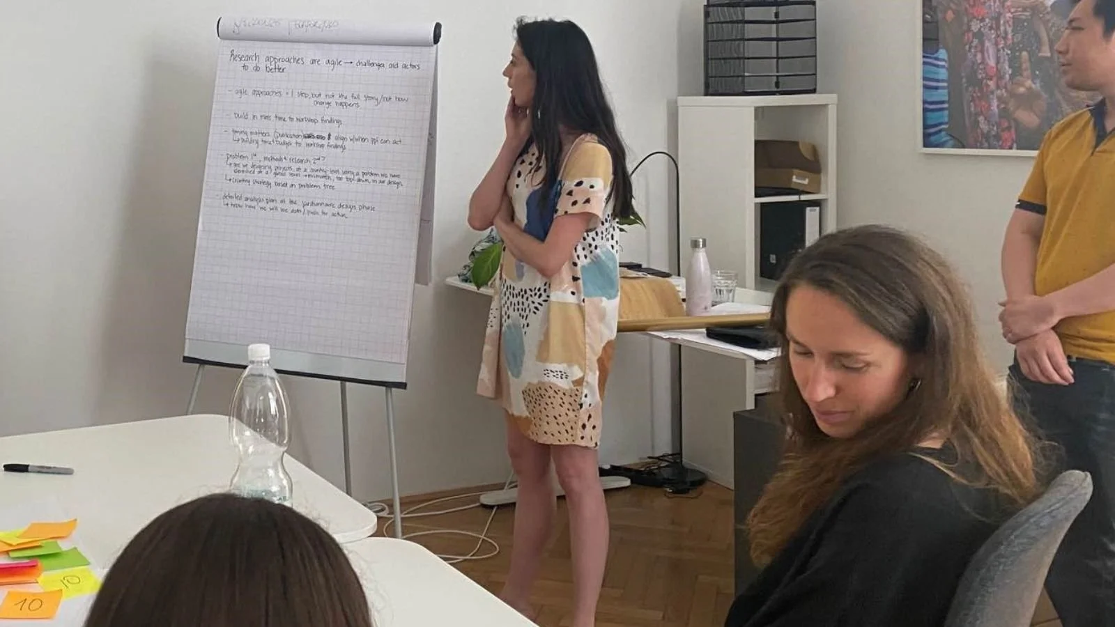 People attending a meeting or workshop in an office, with one woman standing near a flip chart writing notes, and others sitting and listening.
