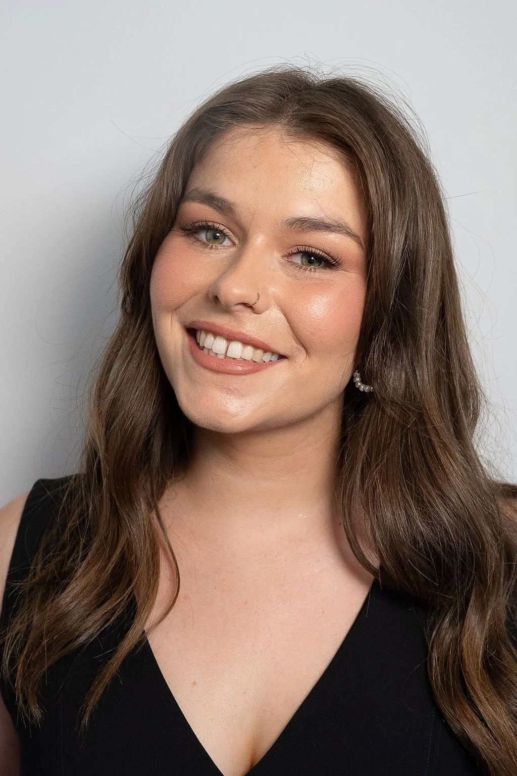 A young woman with long brown hair and natural makeup, wearing a black top, earrings, and a small nose ring, smiling at the camera with a neutral background.