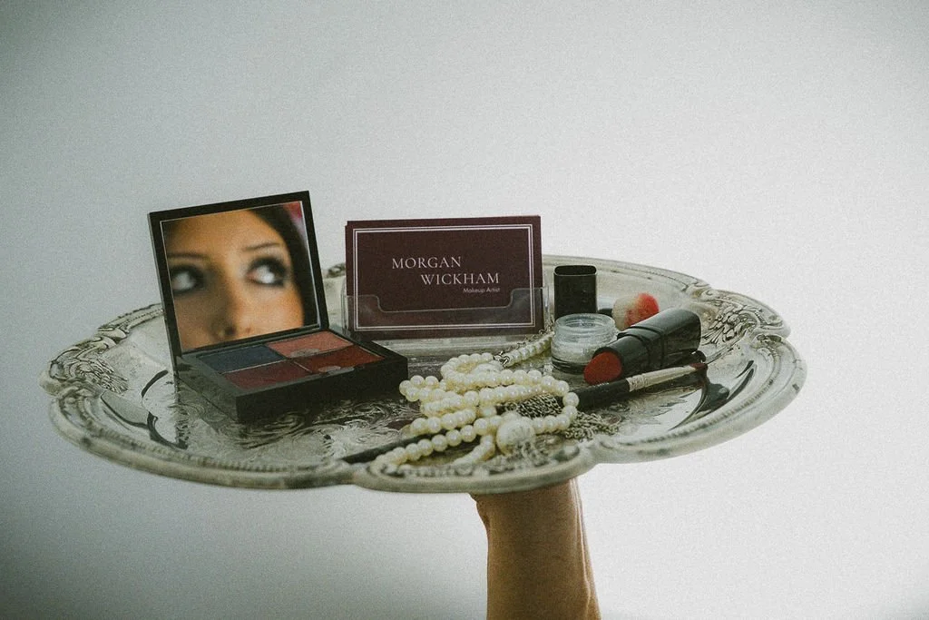 A silver tray on a hand holding various makeup and jewelry items, including a makeup palette, lipstick, beads, and a card that reads 'Morgan Wickham Makeup Artist.'