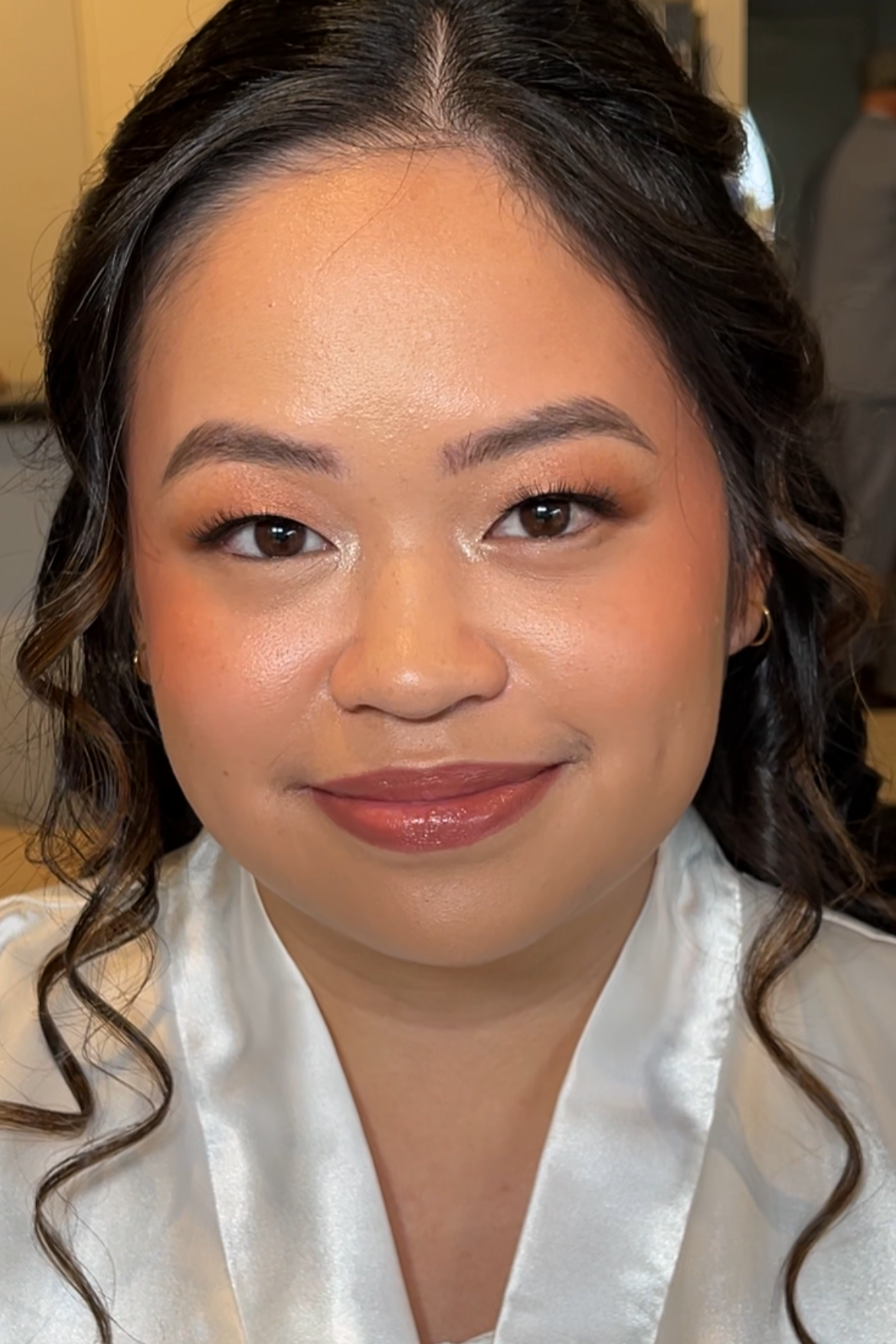 A woman with long dark curly hair wearing makeup with warm toned shimmery eyeshadow, eyeliner, and glossy lipstick, smiling slightly, dressed in a white satin robe on her wedding morning.