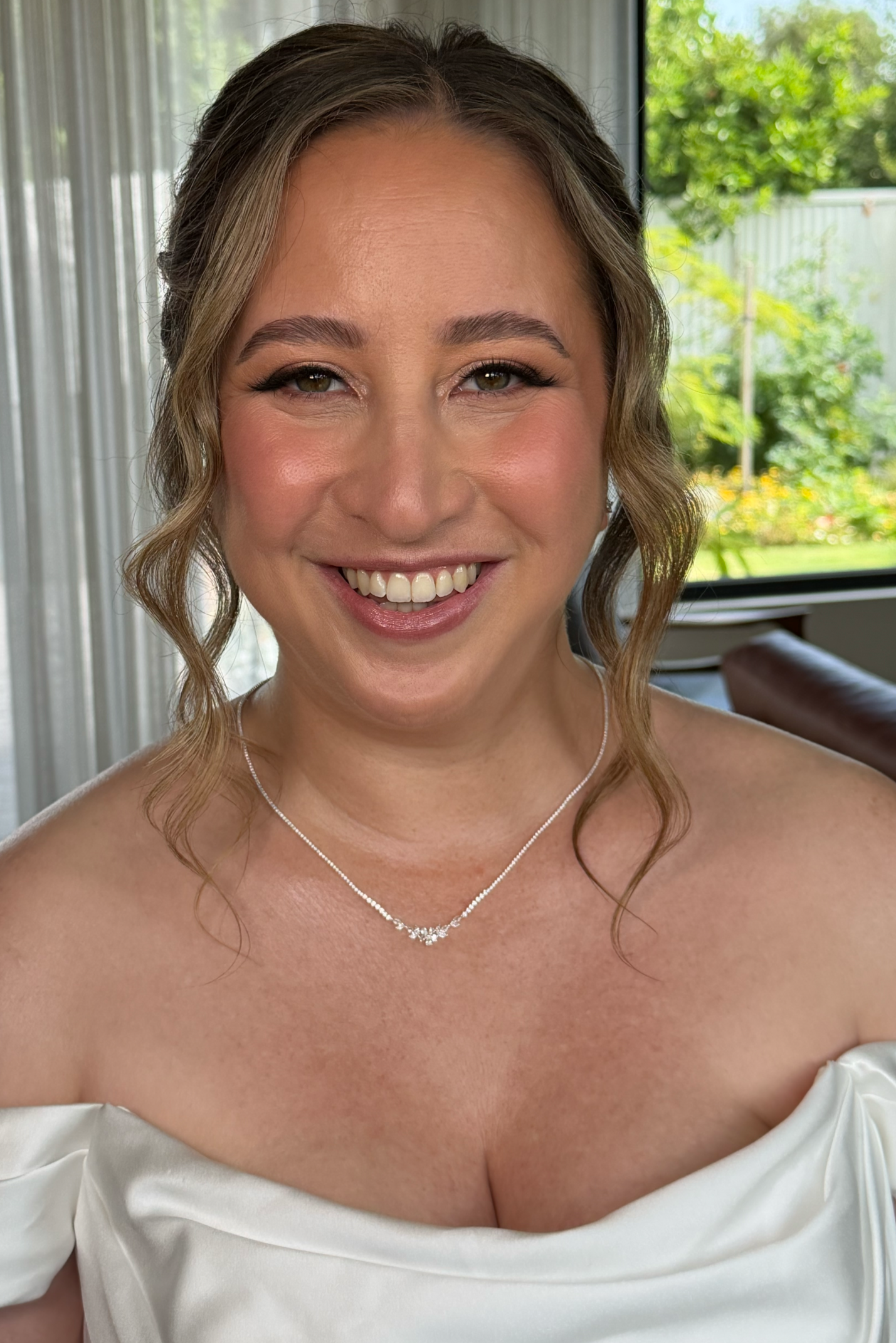 A woman with light brown hair styled in loose waves, wearing glowing bridal makeup, a white off-shoulder wedding dress, and a silver necklace, smiling.