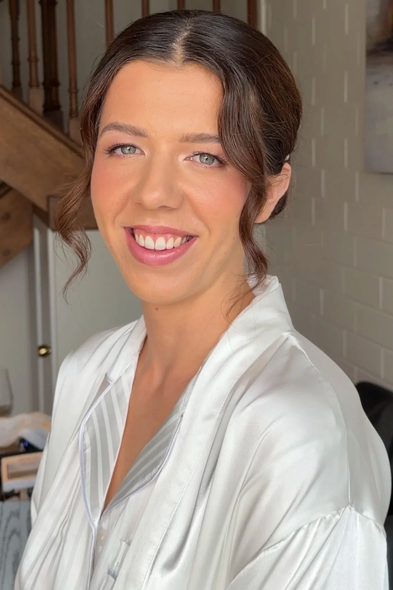 A woman with brown hair styled in a soft up style, blue eyes, wearing light, radiant, pink toned makeup, and a white satin robe, smiling on her wedding morning