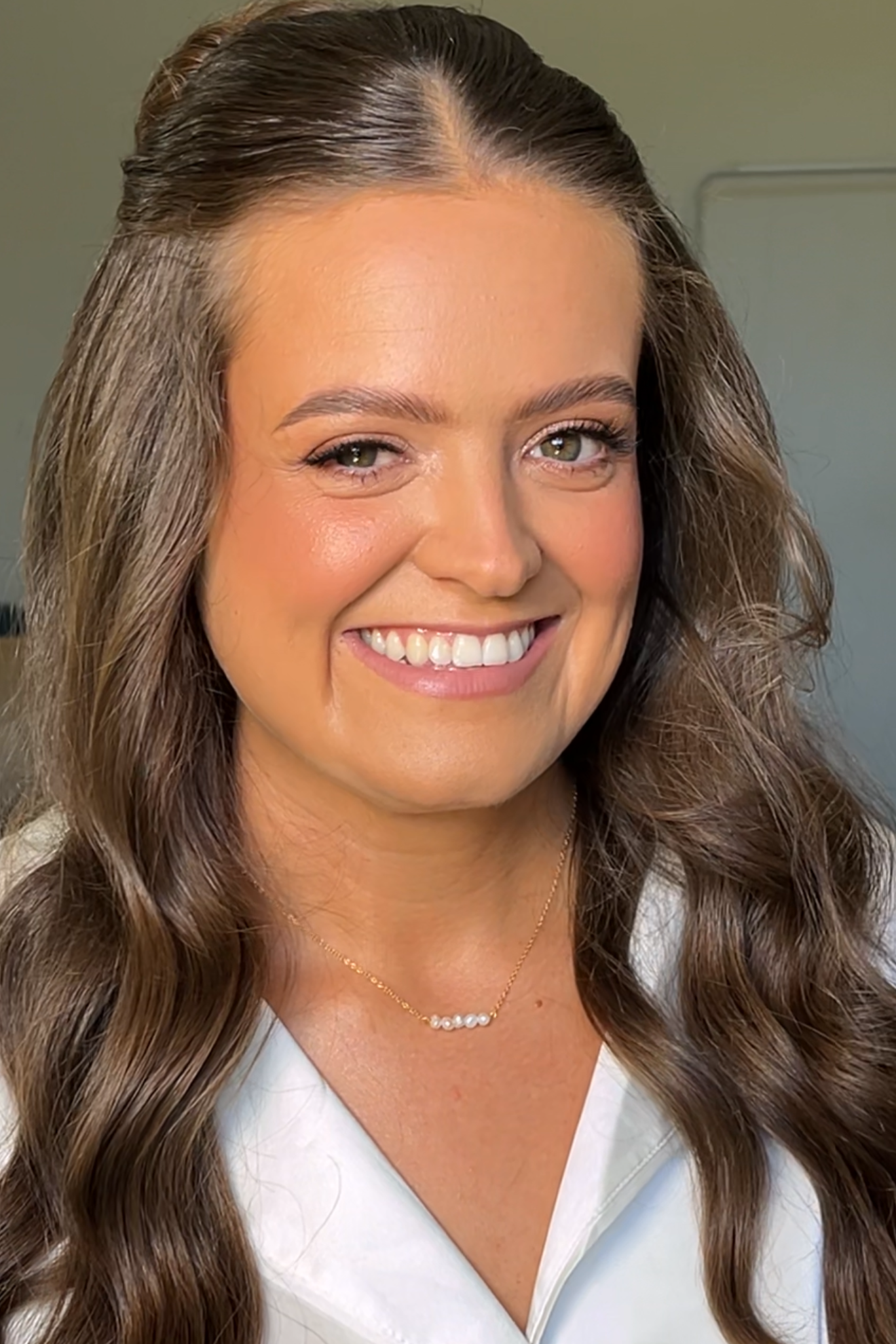 Woman with long, wavy brown hair smiling, wearing beautiful radiant makeup for her wedding, wearing a white top and a delicate pearl necklace.