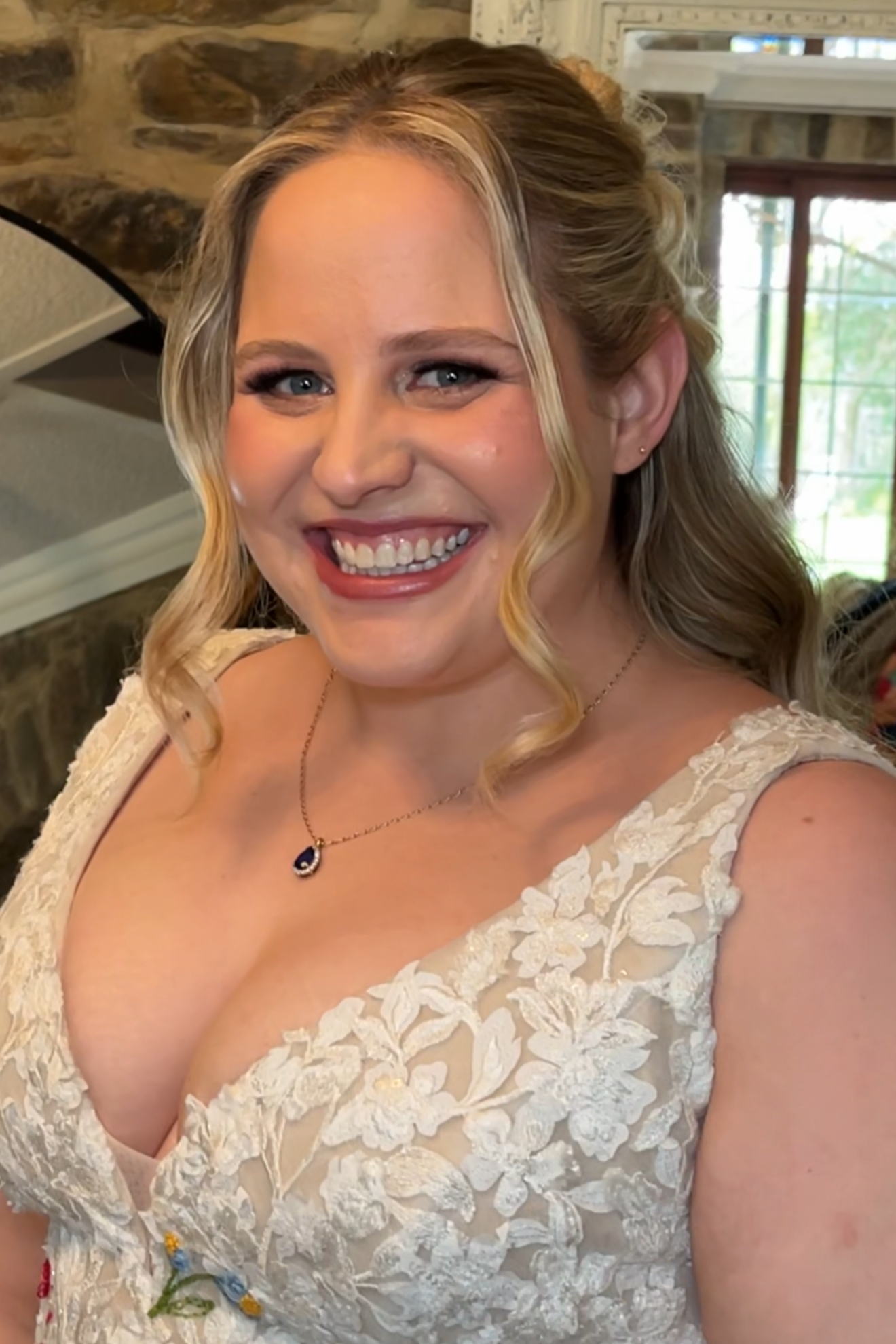 Close-up of a smiling woman with blonde, wavy hair and blue eyes, wearing a cream-colored lace wedding dress with floral embroidery and a small necklace. She has natural glowing makeup.
