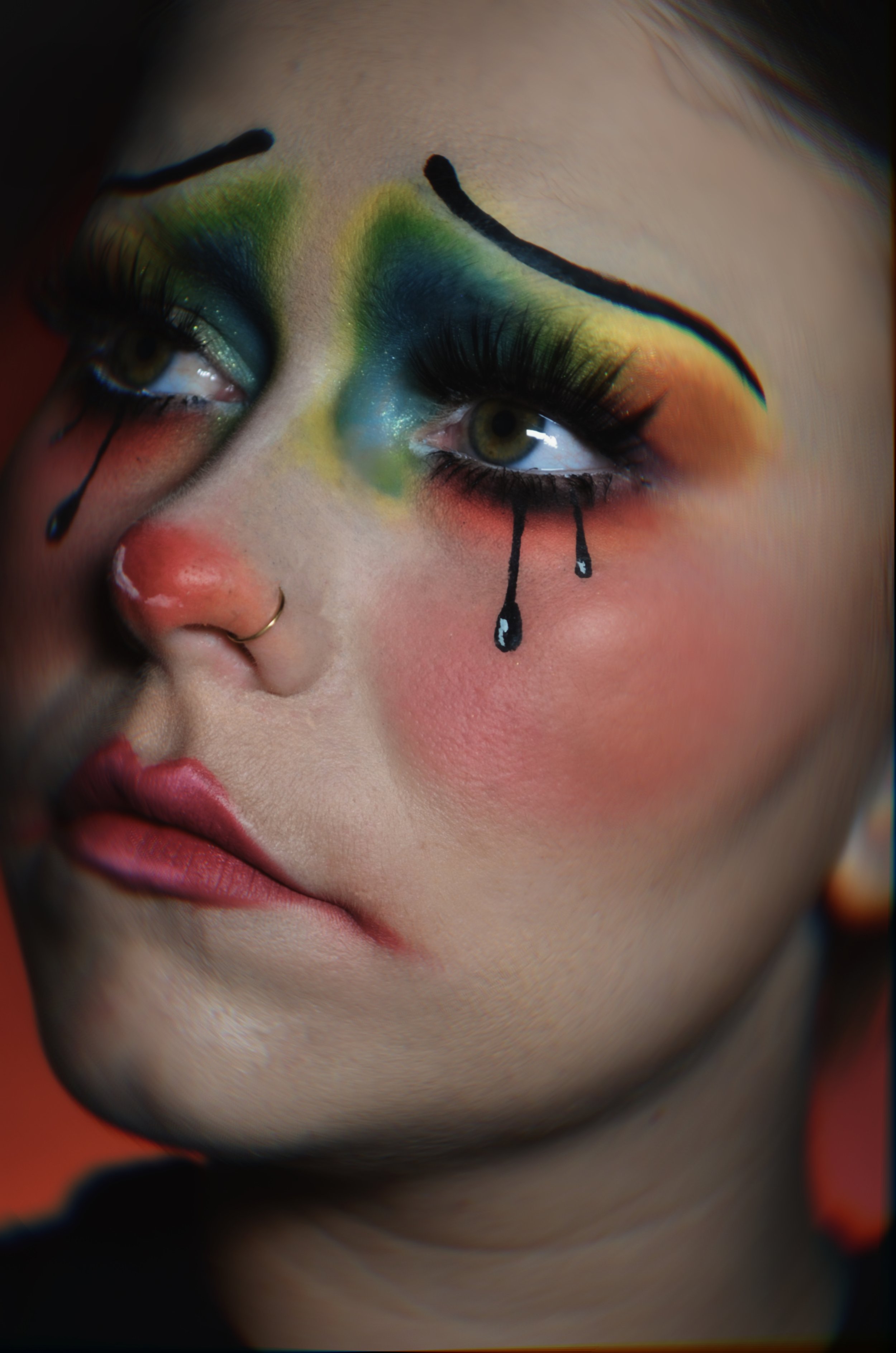 Close-up of a person's face with vibrant, artistic makeup resembling a clown or theatrical style, featuring rainbow eyeshadows, drawn black tears, and exaggerated eyebrows.