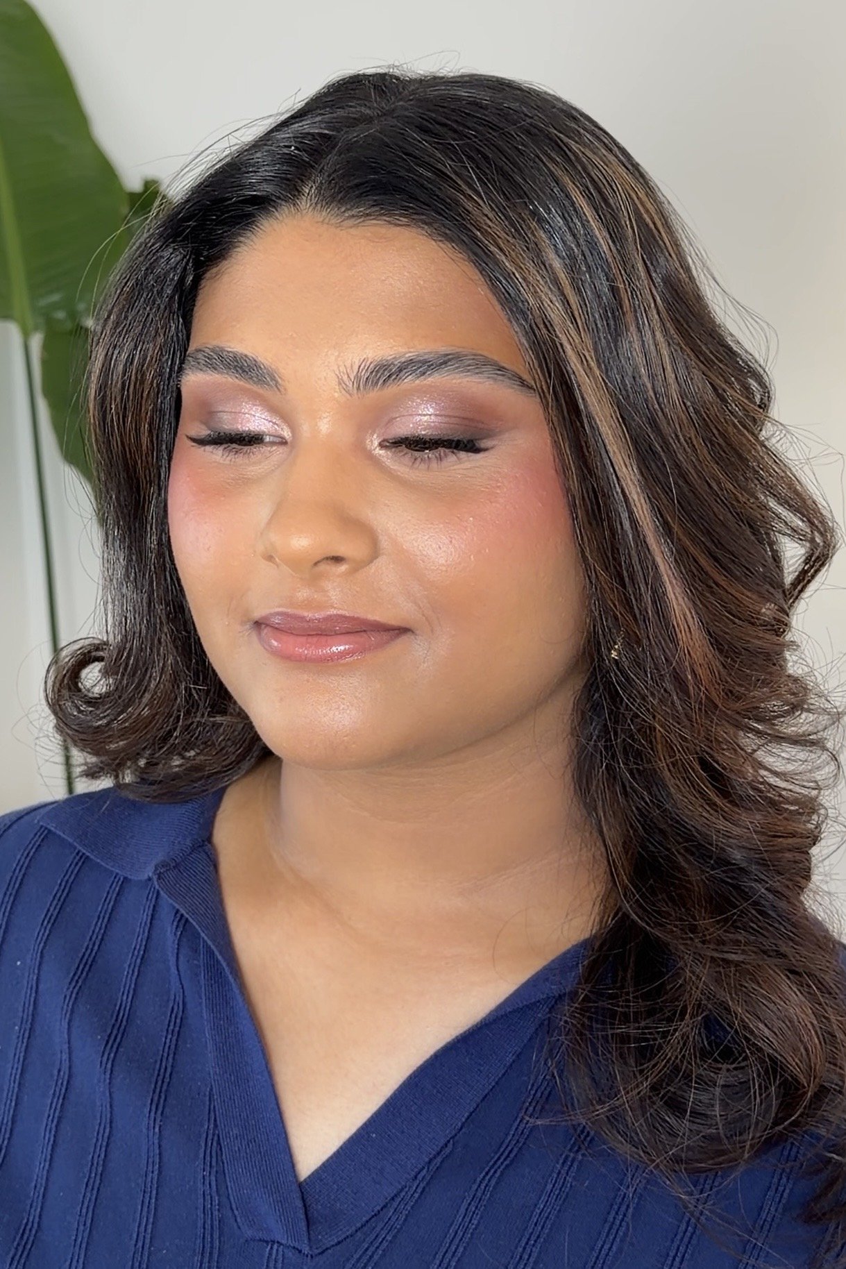 A woman with shoulder-length wavy brown hair with highlights, wearing a blue top, and rose gold toned makeup including eyeshadow, eyeliner, blush, and lipstick, with a neutral expression and closed eyes.