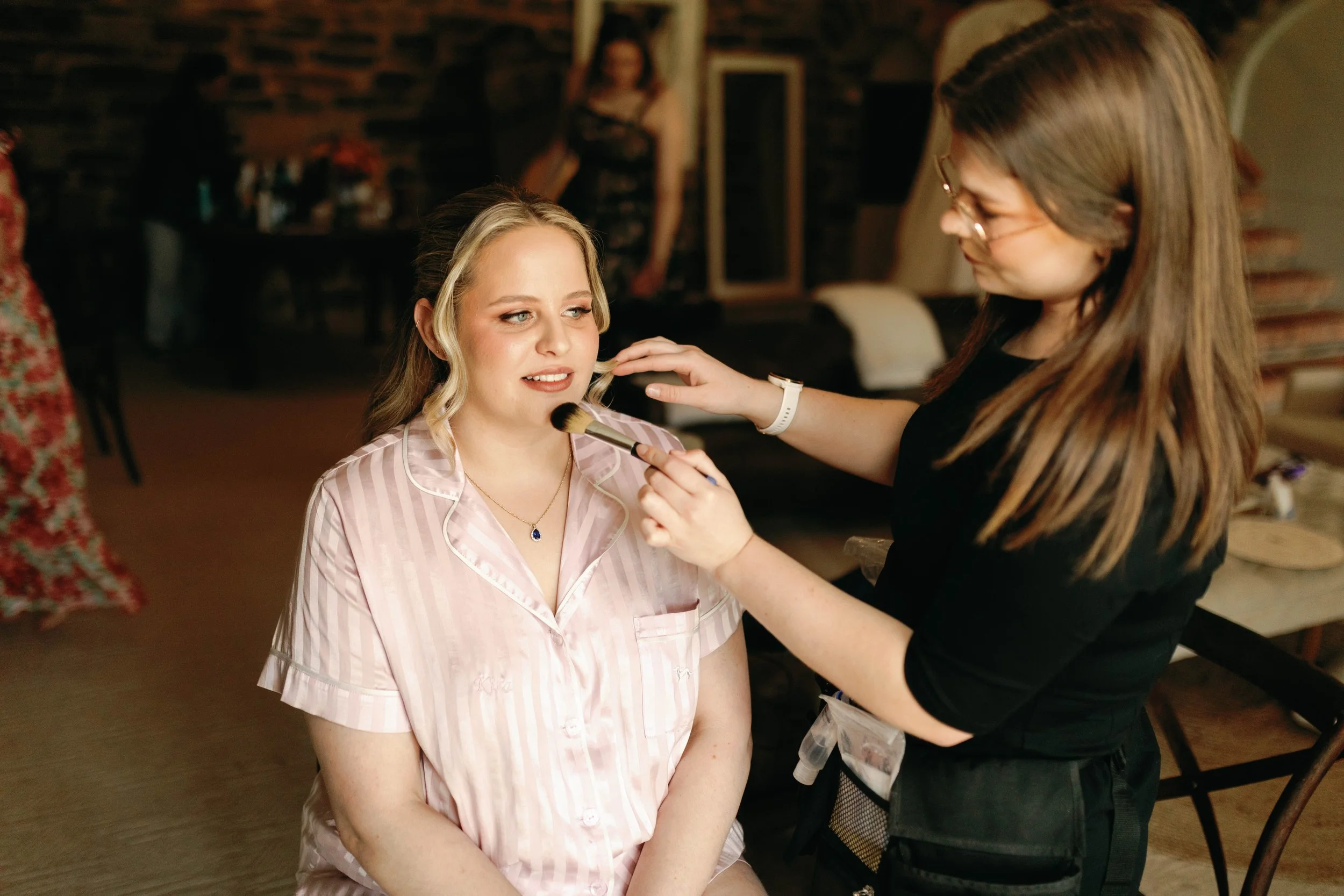 A woman with blonde hair in pink pajamas is getting her makeup done for her wedding by a makeup artist in a cozy indoor setting.
