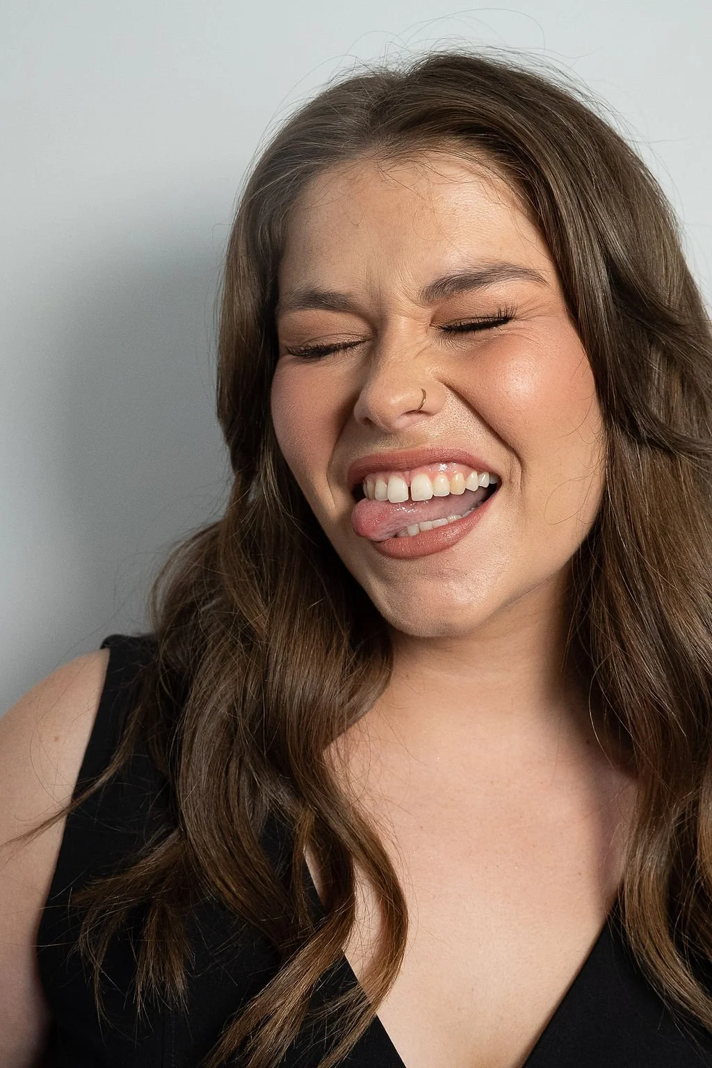 A woman with long brown hair, closed eyes, and a playful expression sticking out her tongue and showing her teeth.