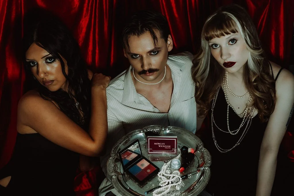 2 women and 1 man dressed in vintage 1920s style, with dramatic makeup and jewelry, gathered around a silver tray with makeup products, pearls, and a name card that reads "Morgan Wickham Makeup Artist", in front of a red velvet curtain.