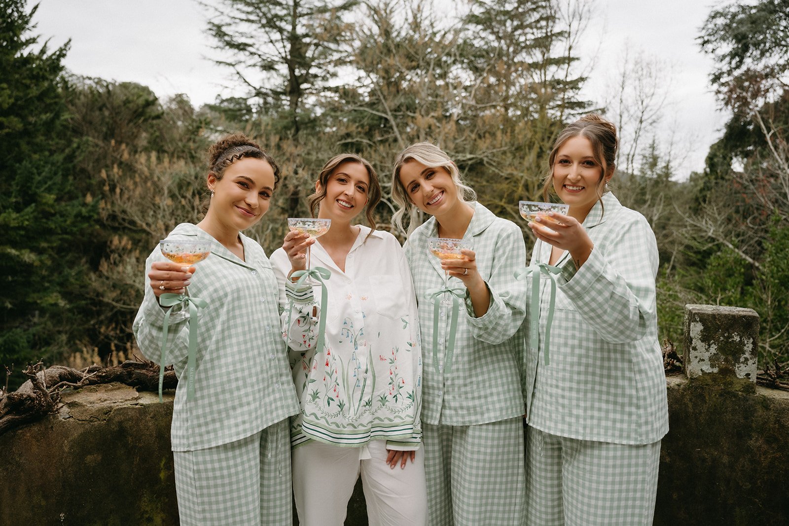Group of five women getting ready for a wedding in pajamas standing outdoors, smiling and holding glasses of champagne.