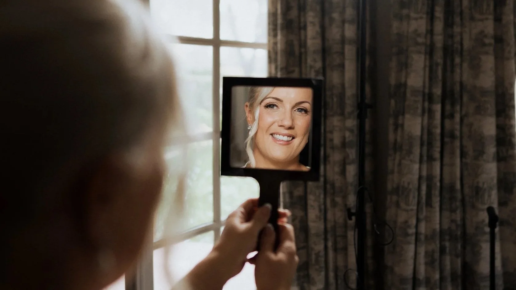 Person holding a mirror reflecting their smiling face looking at her makeup on her wedding day near a window with curtains.