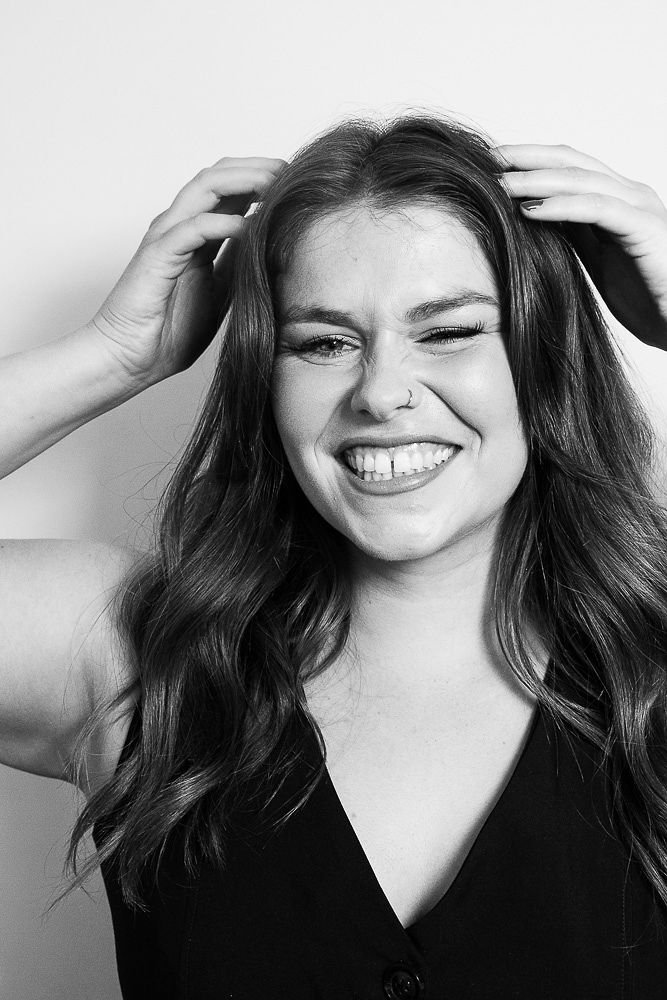 Black and white photo of a woman with wavy hair making a playful face, winking and sticking out her tongue, with hands on her head.