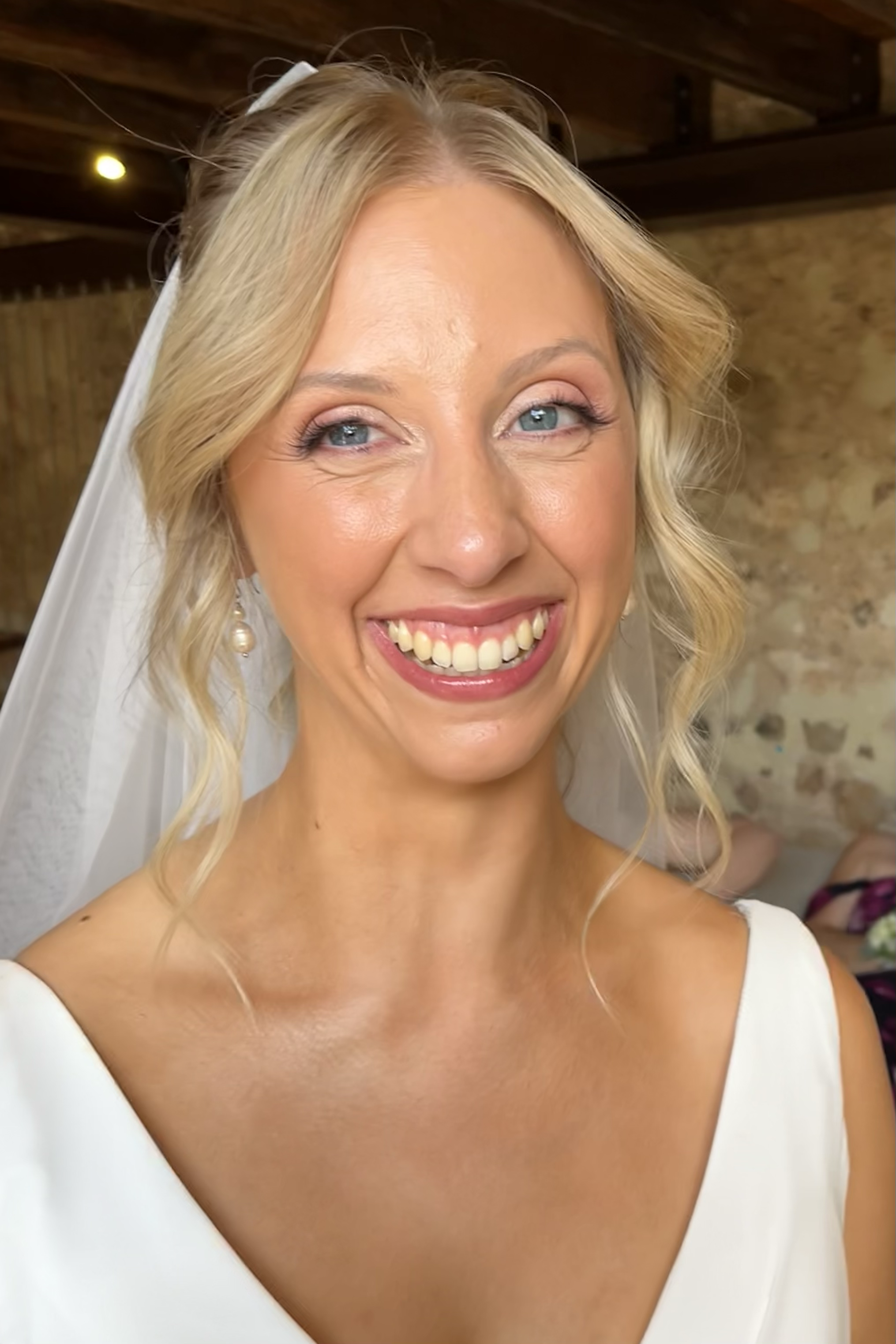 A smiling bride with blonde hair in loose curls and beautiful soft makeup, wearing pearl earrings, a white wedding dress, and a sheer veil, standing indoors with a rustic wooden ceiling and a stone wall background.