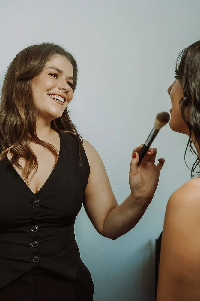A makeup artist applying makeup on a woman with long wavy brown hair, smiling, against a plain light gray background.