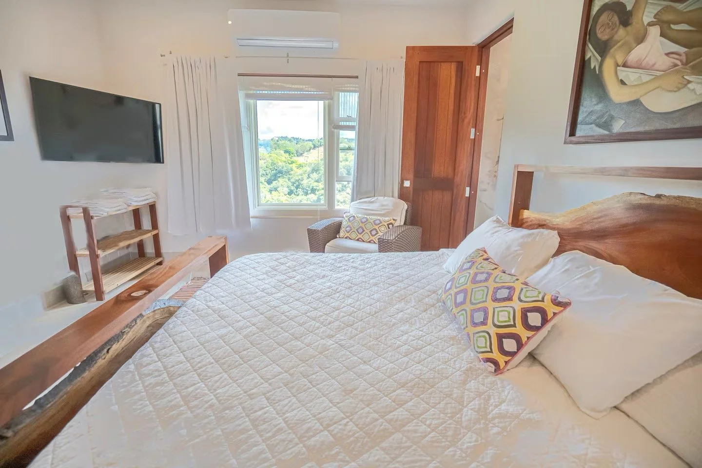 A cozy bedroom with a large wooden bed, cream quilt, and colorful patterned pillows. There's a window with white curtains showing a green landscape outside, a small chair with a patterned pillow near the window, a wall-mounted flat-screen TV, and a w